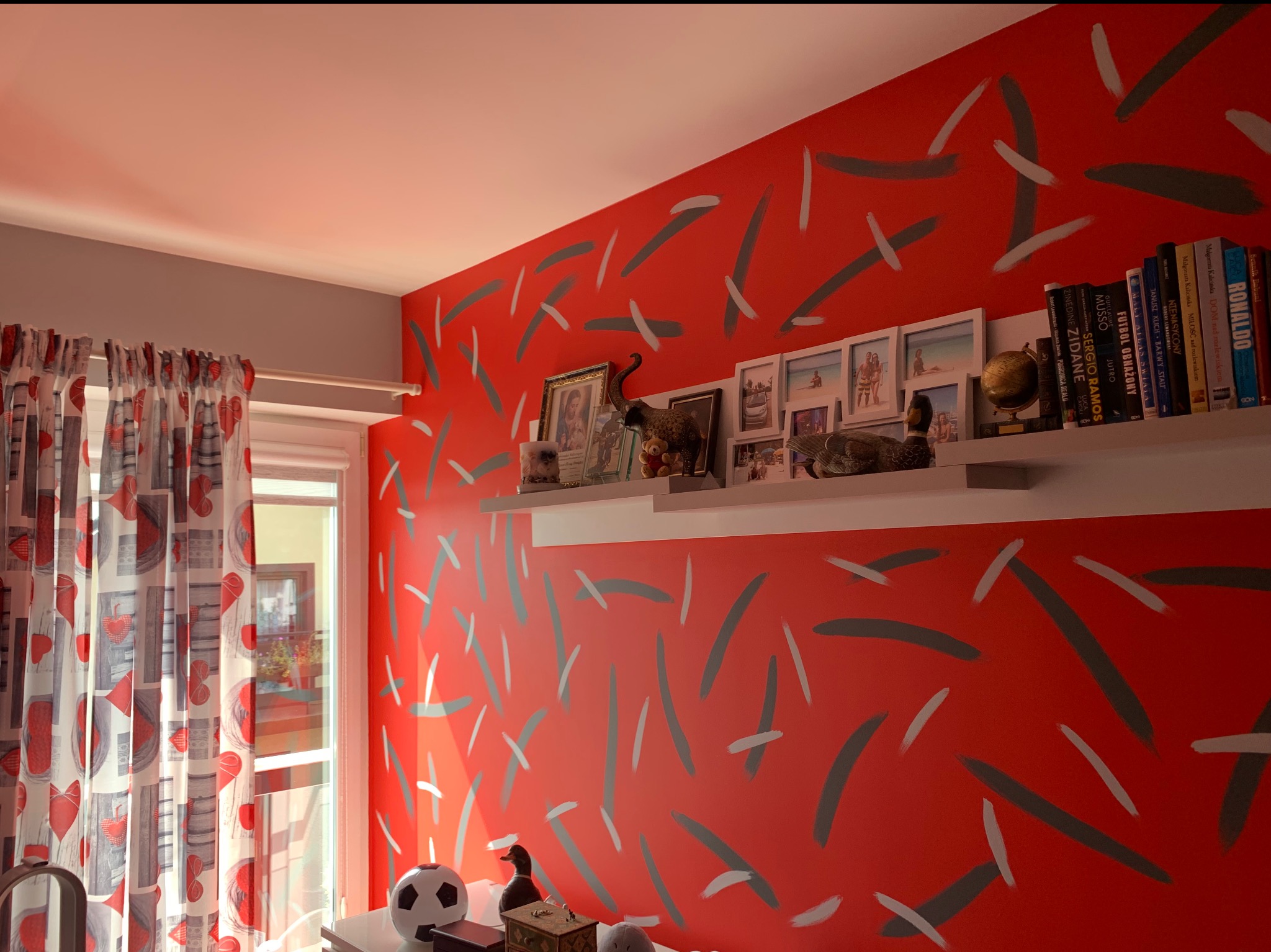 Pokój z czerwoną ścianą ozdobioną szarymi i białymi kreskami, półka z książkami o tematyce piłkarskiej, ramkami ze zdjęciami i dekoracyjnymi figurkami kaczek.