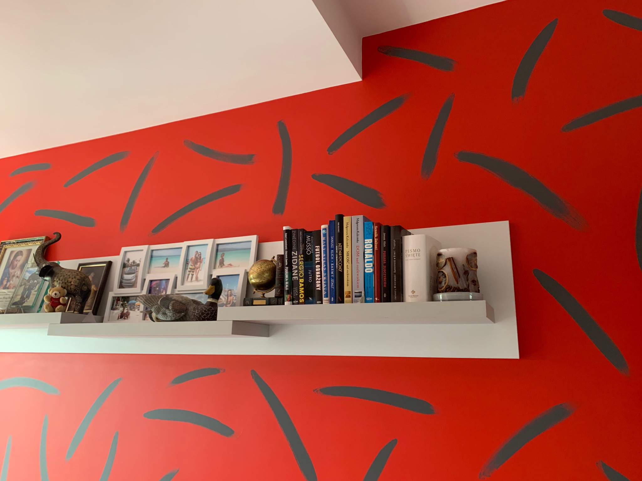 Pomarańczowa ściana z szarymi, nieregularnymi pociągnięciami pędzla, biała półka z książkami, ramkami ze zdjęciami, figurkami i dekoracjami.