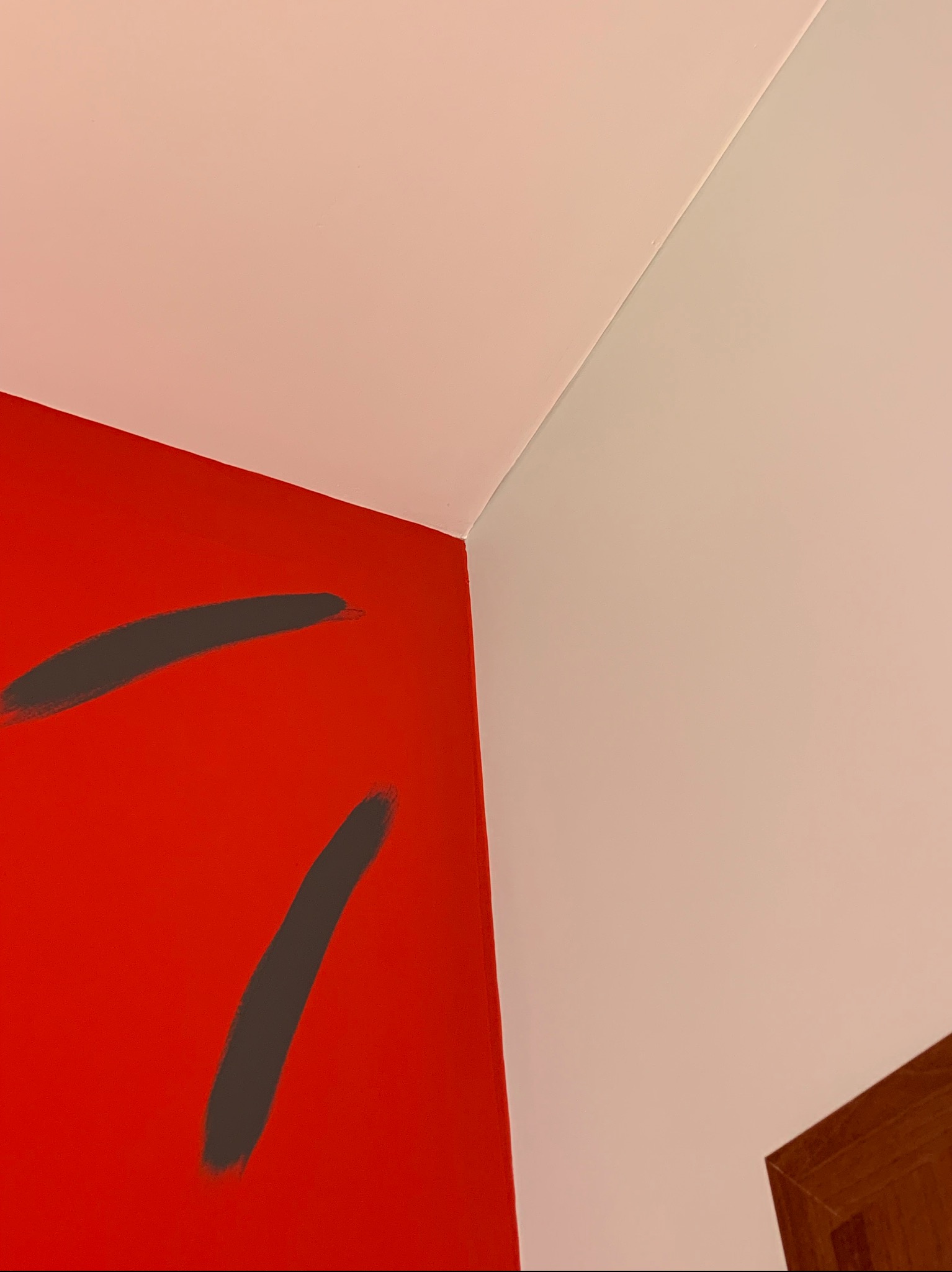 Czerwona ściana z niedomalowanymi szarymi pociągnięciami pędzla w rogu pokoju, widoczny fragment sufitu i sąsiedniej ściany pomalowanej na jasny kolor.