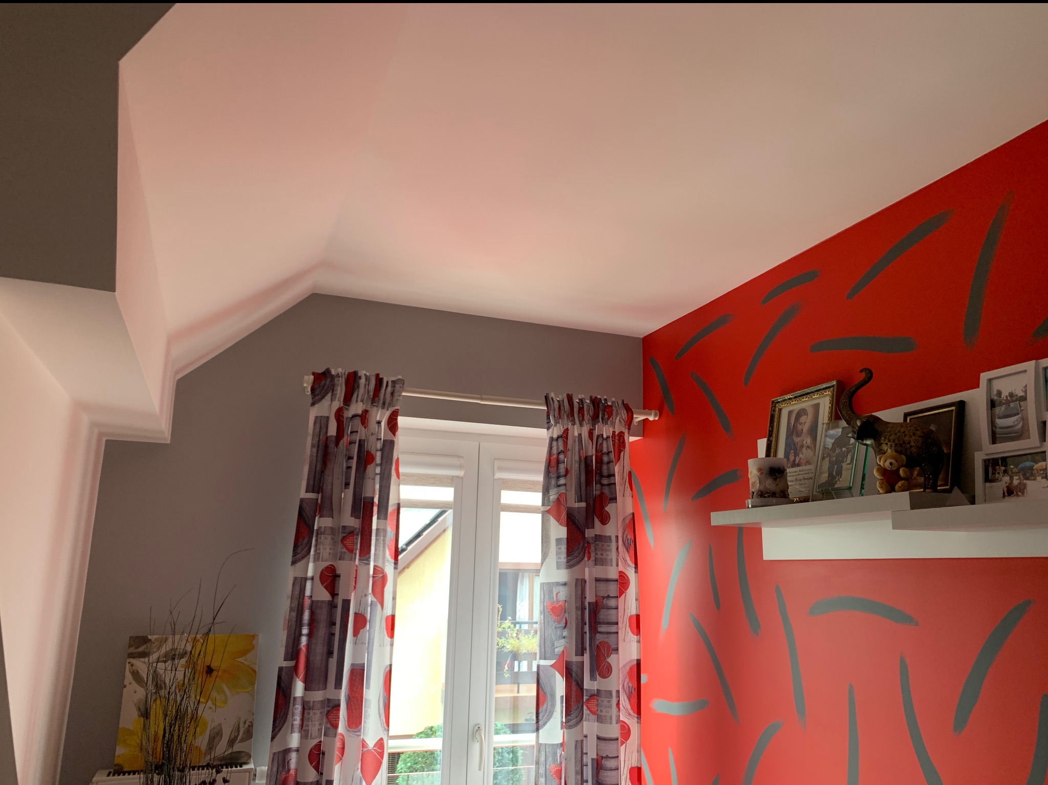 Pokój po odświeżeniu: czerwona ściana z szarymi wzorami, biały sufit, szara ściana boczna, okno z zasłonami w serca, półka z ramkami i dekoracjami.