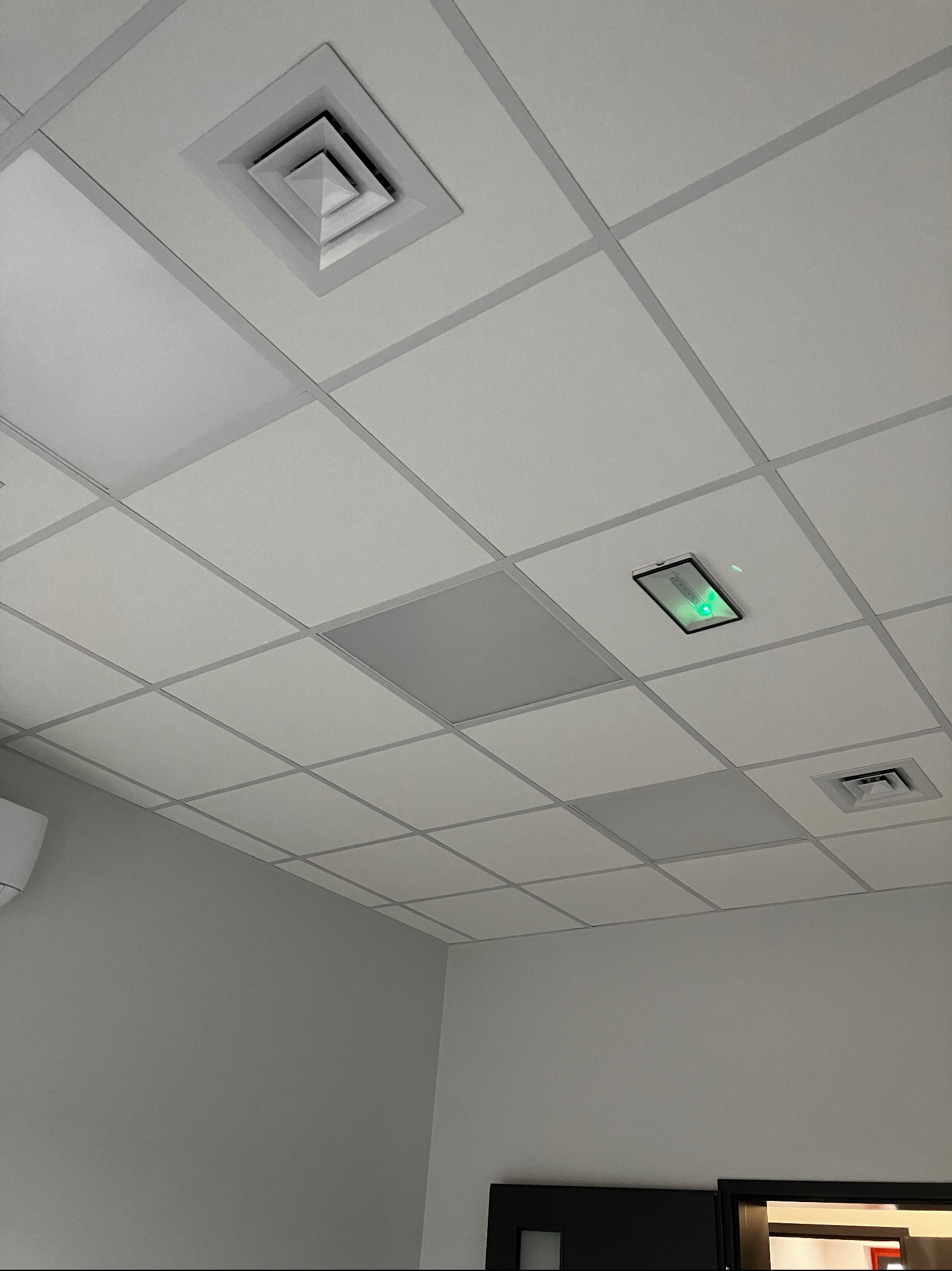 Widok na sufit podwieszany z kratką wentylacyjną, panelem oświetleniowym LED z zielonym światłem i panelem rewizyjnym w biurze o szarych ścianach.