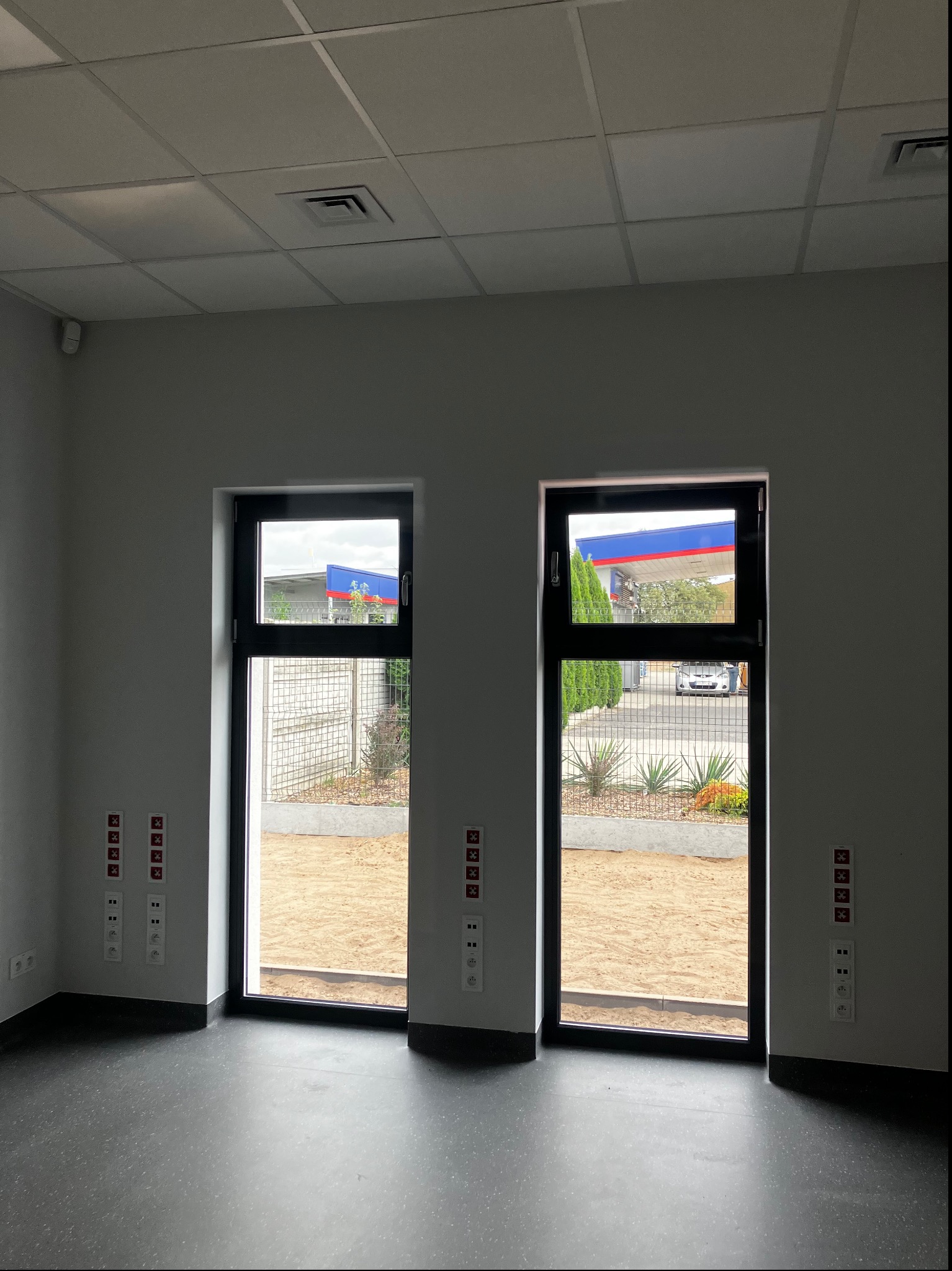 Wnętrze nowo wykończonego pomieszczenia z widocznymi dwoma oknami, przez które widać stację benzynową, na ścianach rozmieszczone gniazdka elektryczne i włączniki z charakterystycznymi czerwonymi...