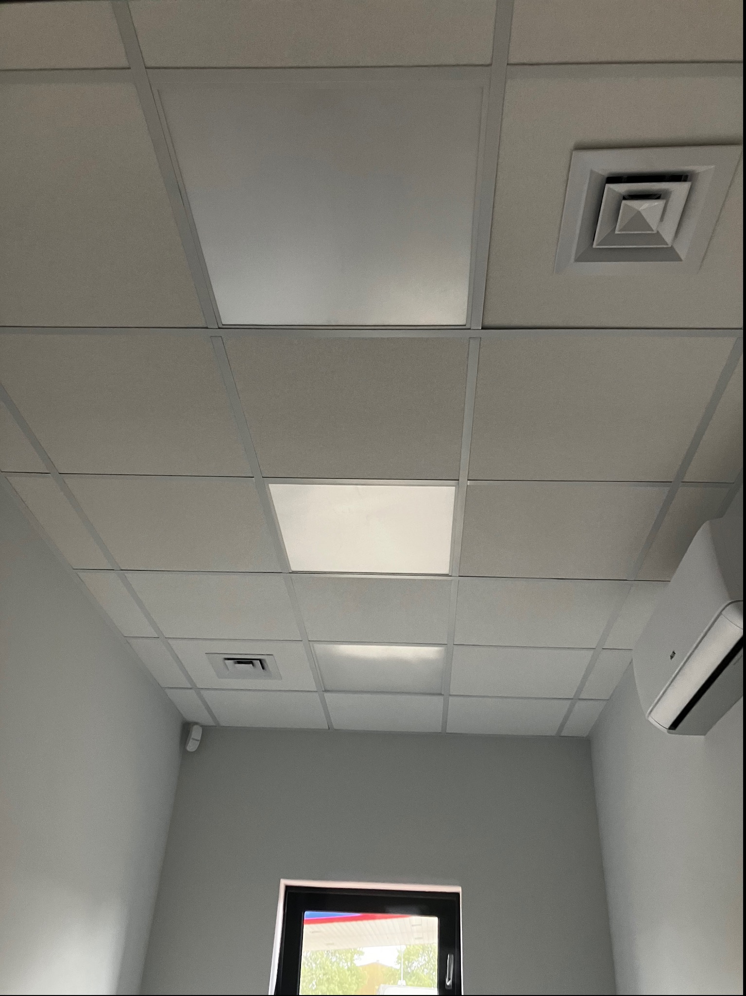 Kasetonowy sufit podwieszany z rastrowym oświetleniem LED, kratka wentylacyjna i klimatyzator ścienny w jasnym pomieszczeniu biurowym.