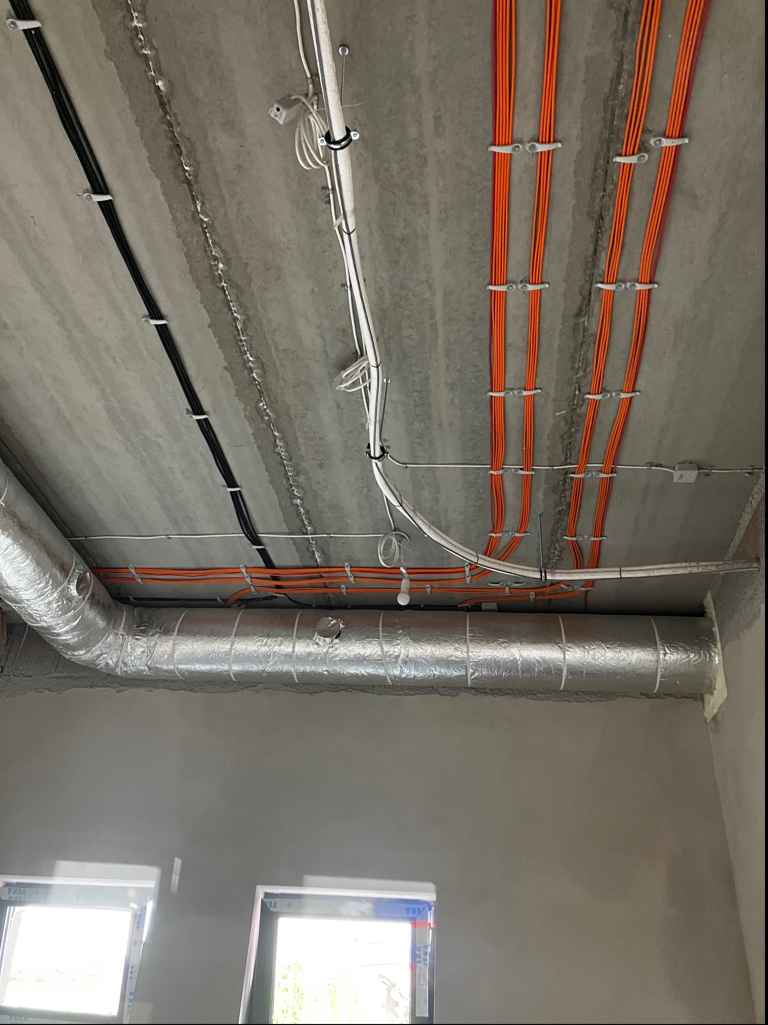 Surowe wnętrze w trakcie wykańczania: widoczne przewody elektryczne w kolorze pomarańczowym i czarnym, przymocowane do betonowego sufitu za pomocą białych uchwytów, oraz srebrny kanał wentylacyjny...