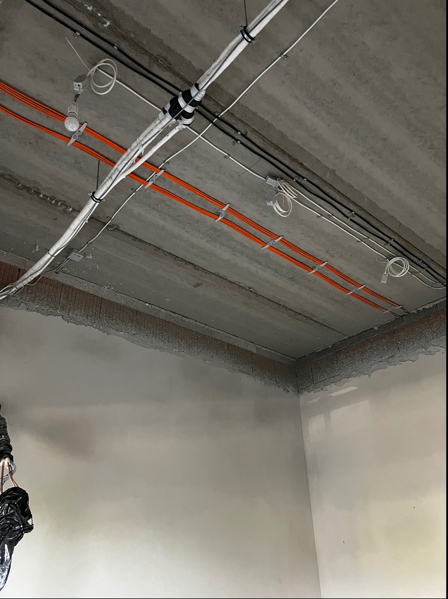 Surowy betonowy sufit z widocznymi przewodami elektrycznymi w kolorach pomarańczowym, białym i czarnym, przymocowanymi uchwytami; ściany w trakcie wykończenia.