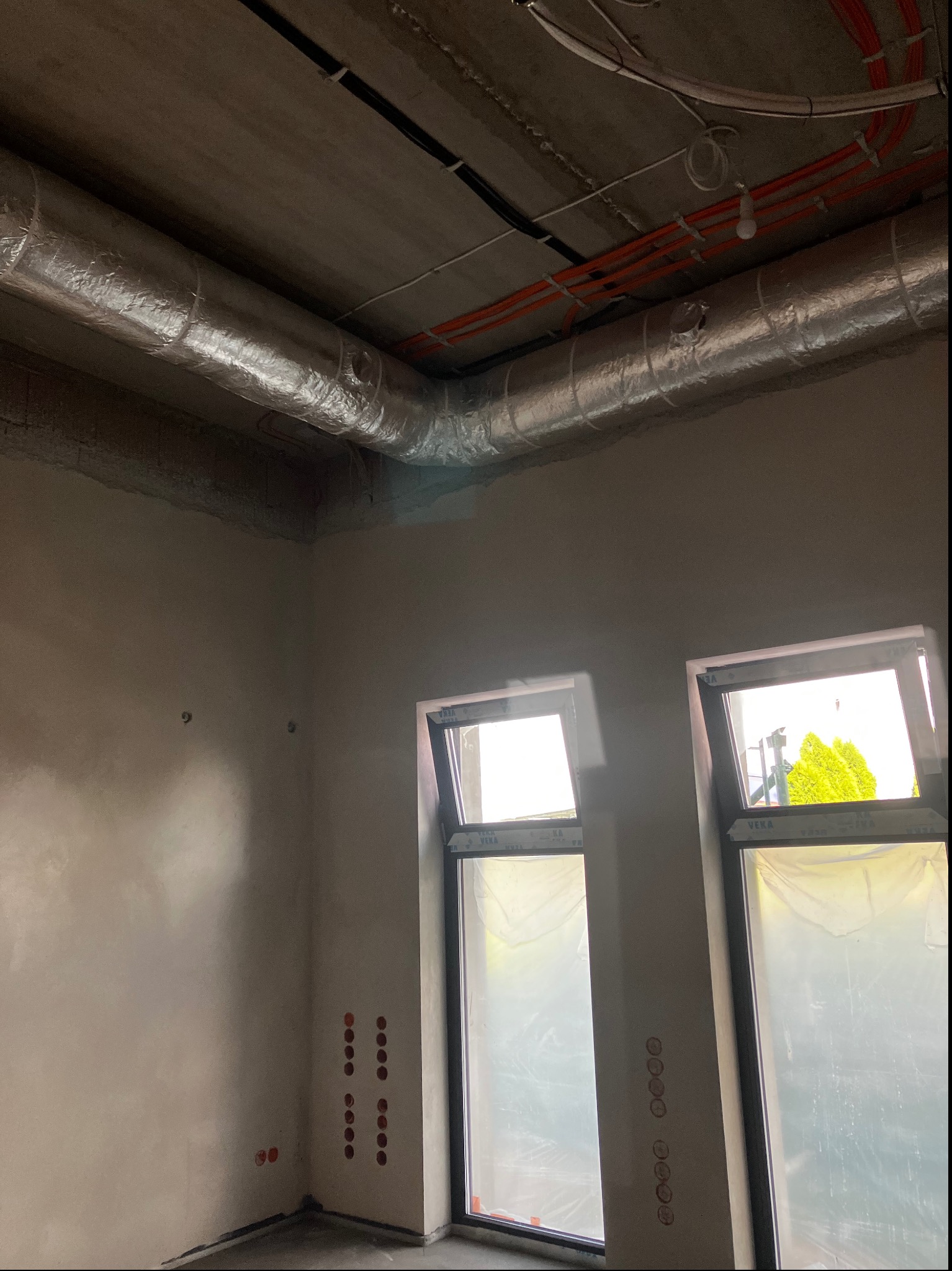 Surowy stan wnętrza z widocznymi przewodami elektrycznymi pod sufitem, rurami wentylacyjnymi w izolacji oraz przygotowanymi punktami elektrycznymi na ścianach, dwa okna zabezpieczone folią ochronną.