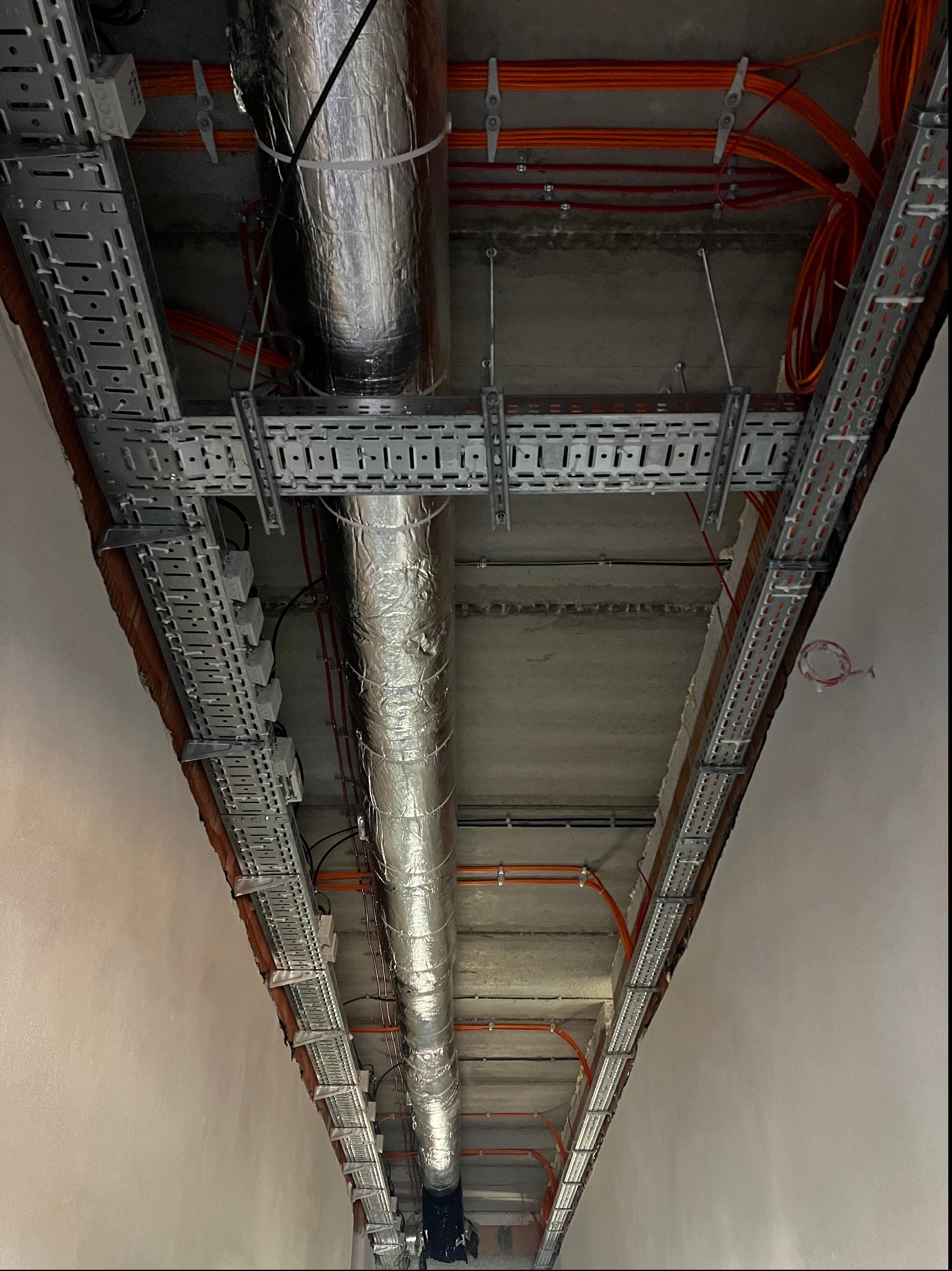 Widok sufitu z zamontowanymi korytkami kablowymi, rurami wentylacyjnymi owiniętymi folią aluminiową oraz przewodami elektrycznymi w kolorze pomarańczowym, mocowanymi do sufitu za pomocą uchwytów.