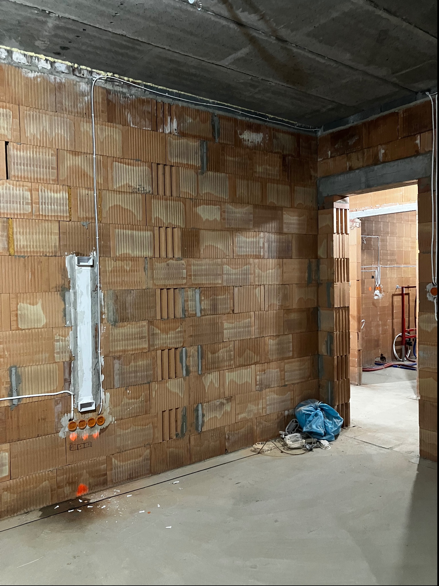 Surowy stan wnętrza z widocznymi przewodami elektrycznymi poprowadzonymi po ścianie z cegły ceramicznej, gniazdka elektryczne zamontowane w puszce, niebieski worek z odpadami budowlanymi na podłodze.