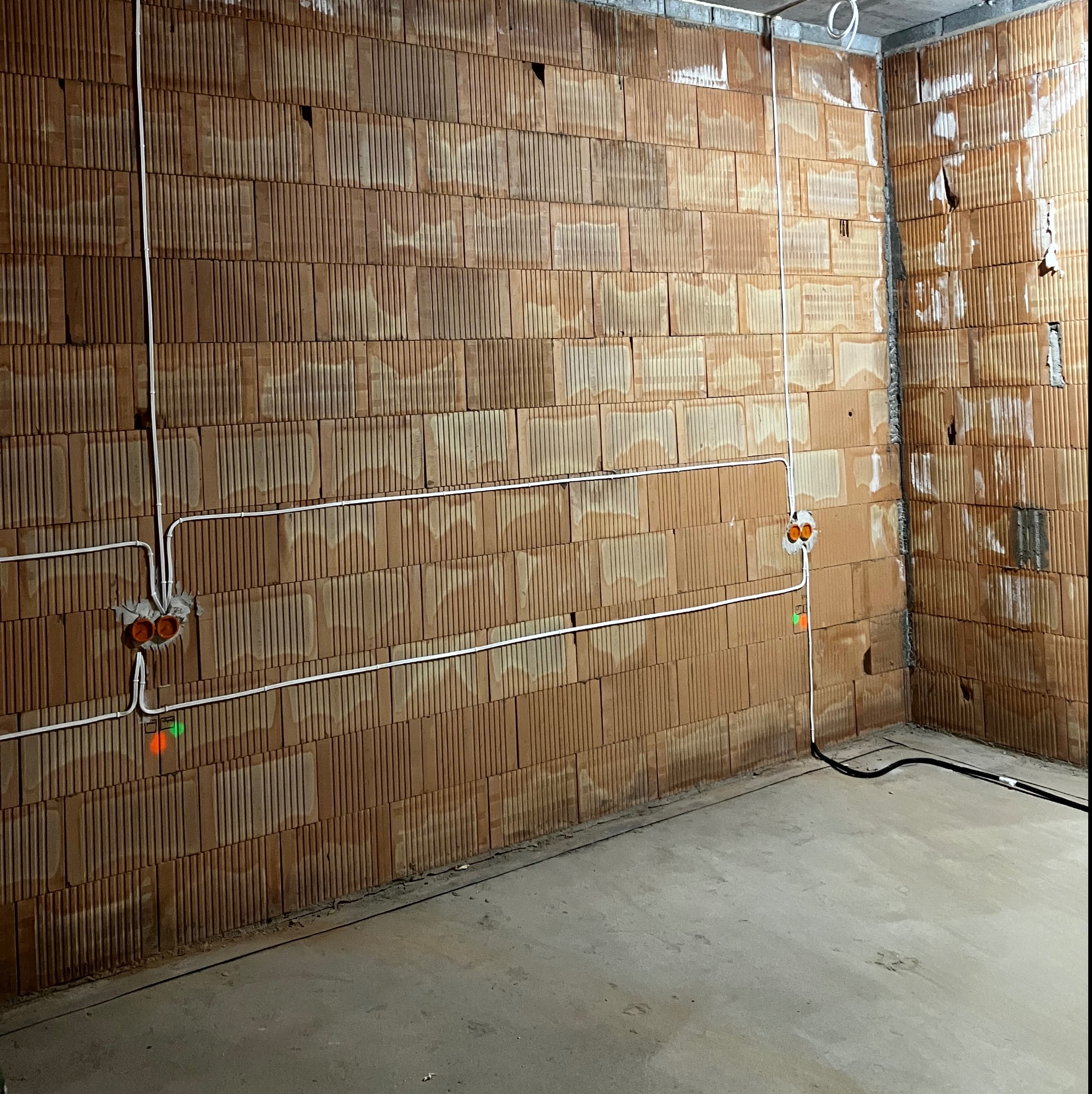 Świeżo położone przewody elektryczne w białych rurkach na ścianie z cegieł dziurawki, widoczne pomarańczowe puszki pod gniazdka i kable na podłodze w surowym stanie.