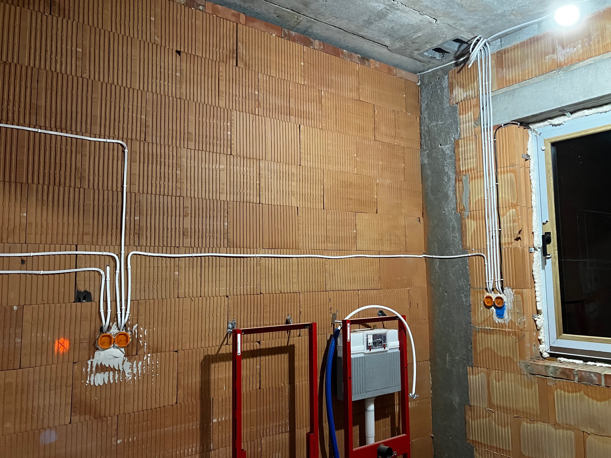 Wewnętrzna instalacja elektryczna w surowym stanie: przewody w peszlach prowadzone po ścianie z pustaków ceramicznych, puszki elektryczne w tynku, widoczne elementy instalacji sanitarnej w tle.