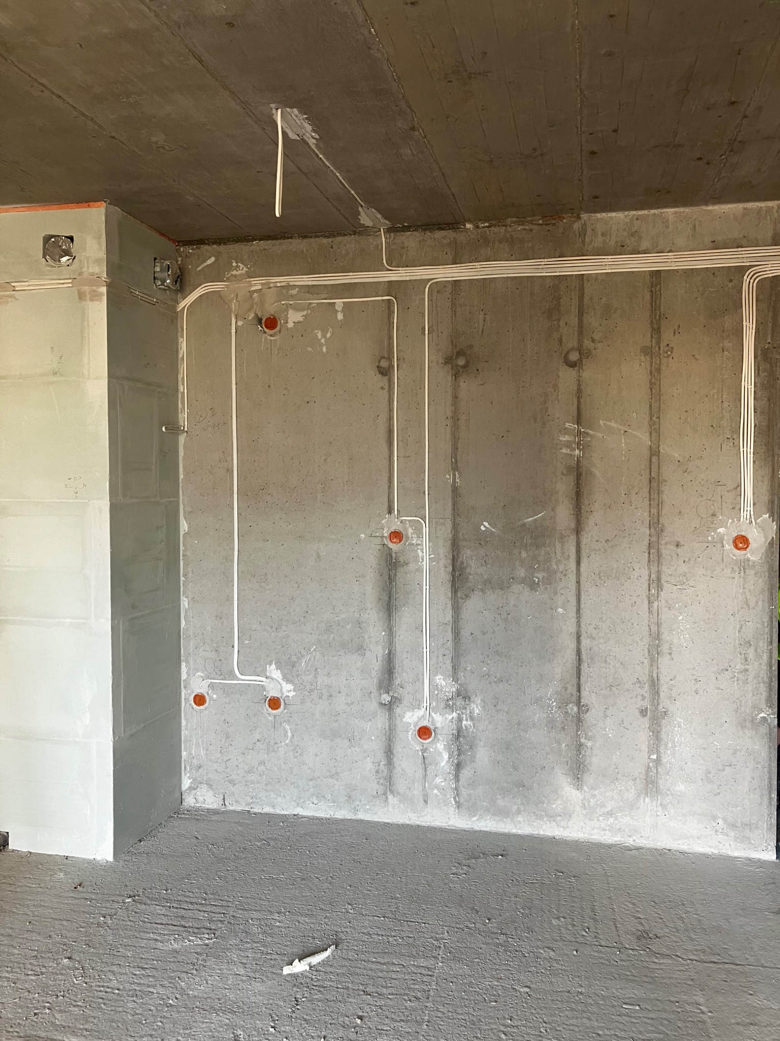 Surowy betonowy pokój w trakcie instalacji elektrycznej: białe kable prowadzone po ścianach i suficie do pomarańczowych puszek, z fragmentem ściany obudowanej płytami gipsowymi po lewej stronie.