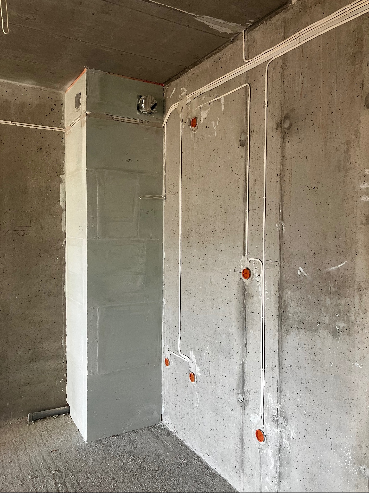 Surowa betonowa ściana z widocznymi przewodami elektrycznymi w białych listwach oraz pomarańczowymi puszkami instalacyjnymi, obok pionowy szyb wentylacyjny z wykończeniem gipsowym przed malowaniem.