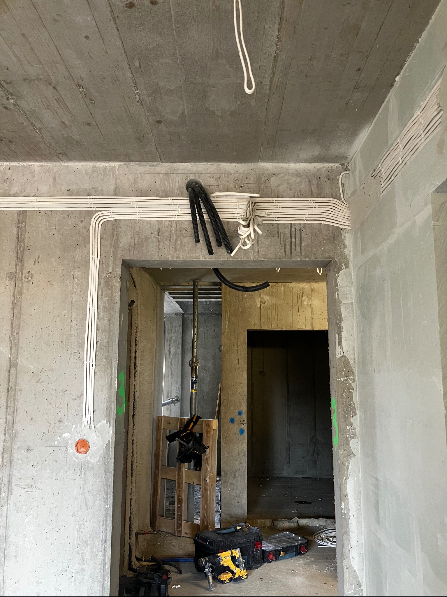 Surowy stan wnętrza w trakcie prac elektrycznych: widoczne kable prowadzone po betonowym suficie i ścianach, skrzynki narzędziowe DeWalt na podłodze, podpora budowlana w przejściu między...