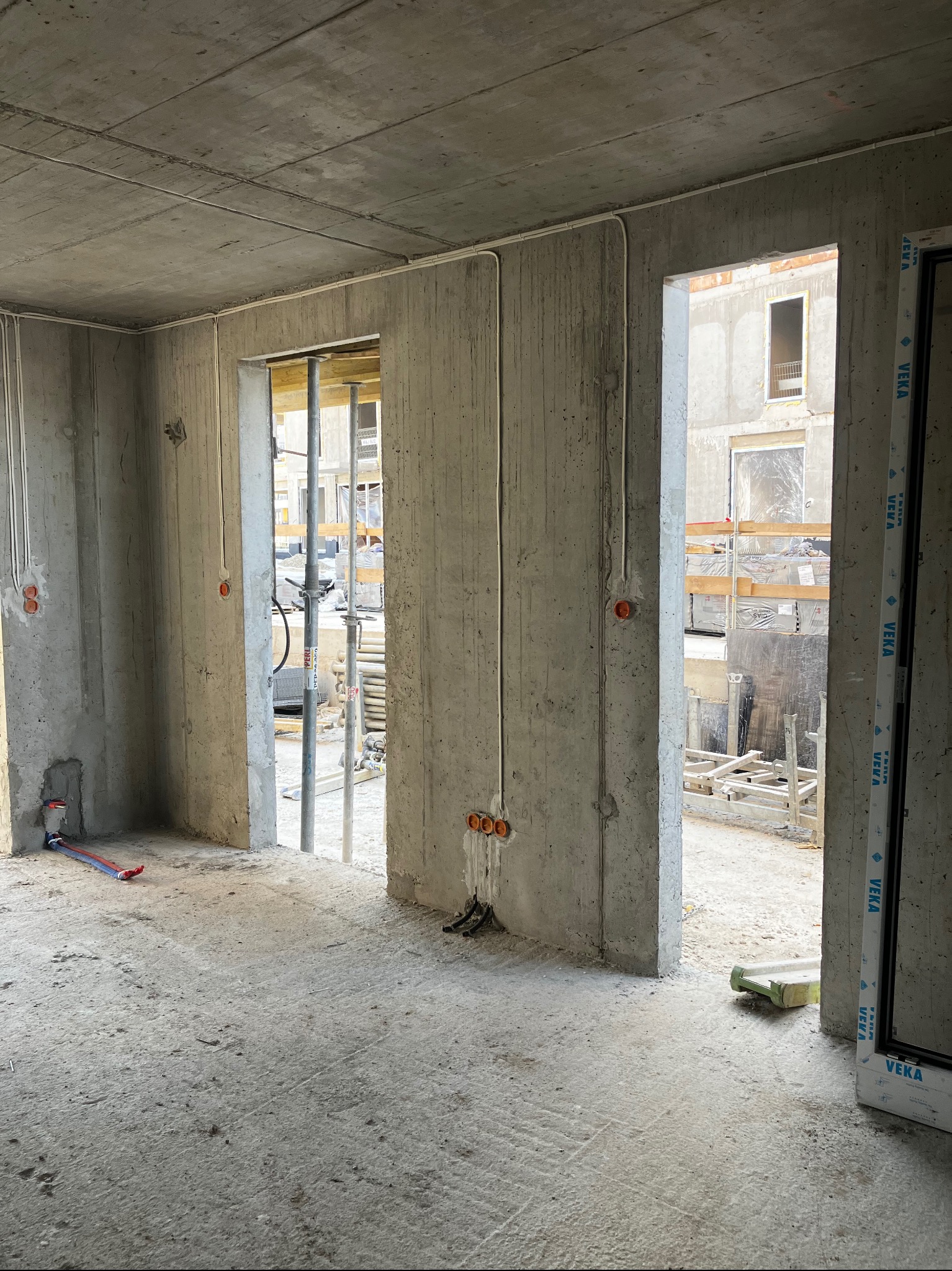 Surowe wnętrze w stanie deweloperskim z widocznymi przewodami elektrycznymi w peszlach, instalacją wodną i otworami pod gniazdka w ścianach, przez które widać rusztowania na zewnątrz.