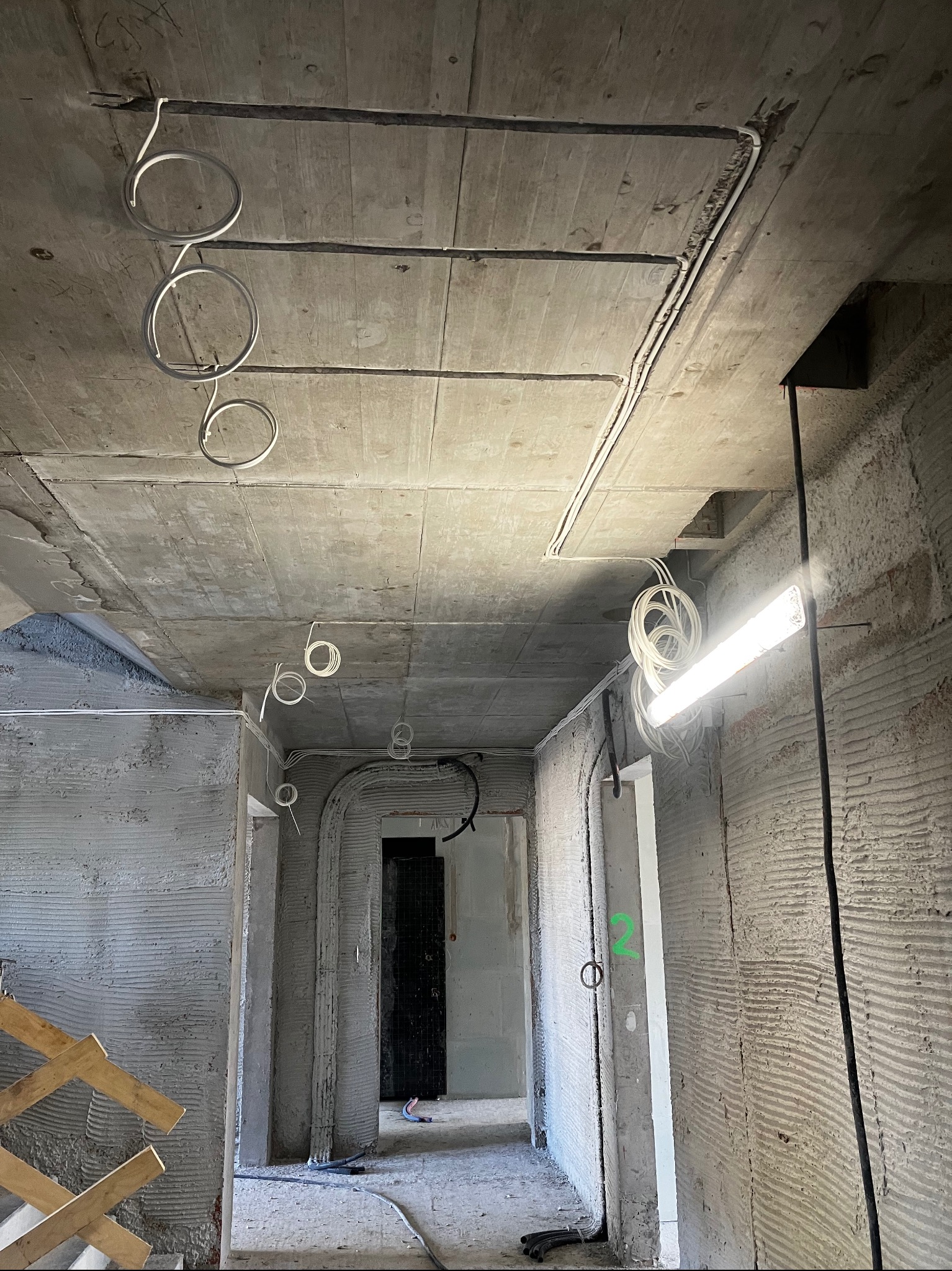 Surowy betonowy sufit z widocznymi kablami elektrycznymi zwisającymi w pętlach, surowe ściany w trakcie wykończenia, tymczasowe oświetlenie budowlane.