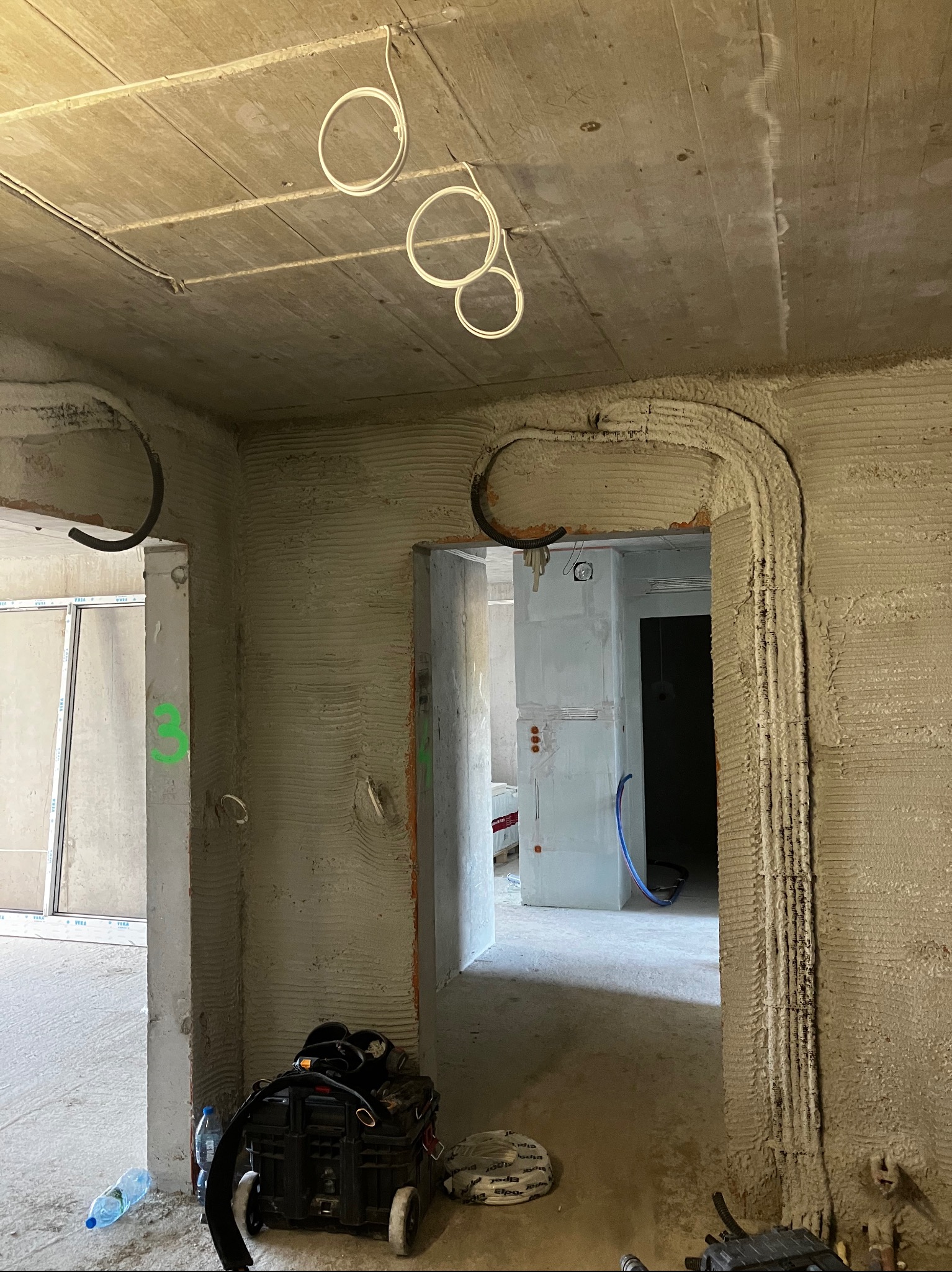 Wnętrze w stanie surowym z widocznymi przewodami elektrycznymi ułożonymi pod tynkiem na ścianach i suficie, narzędzia elektryczne w skrzynce na podłodze.