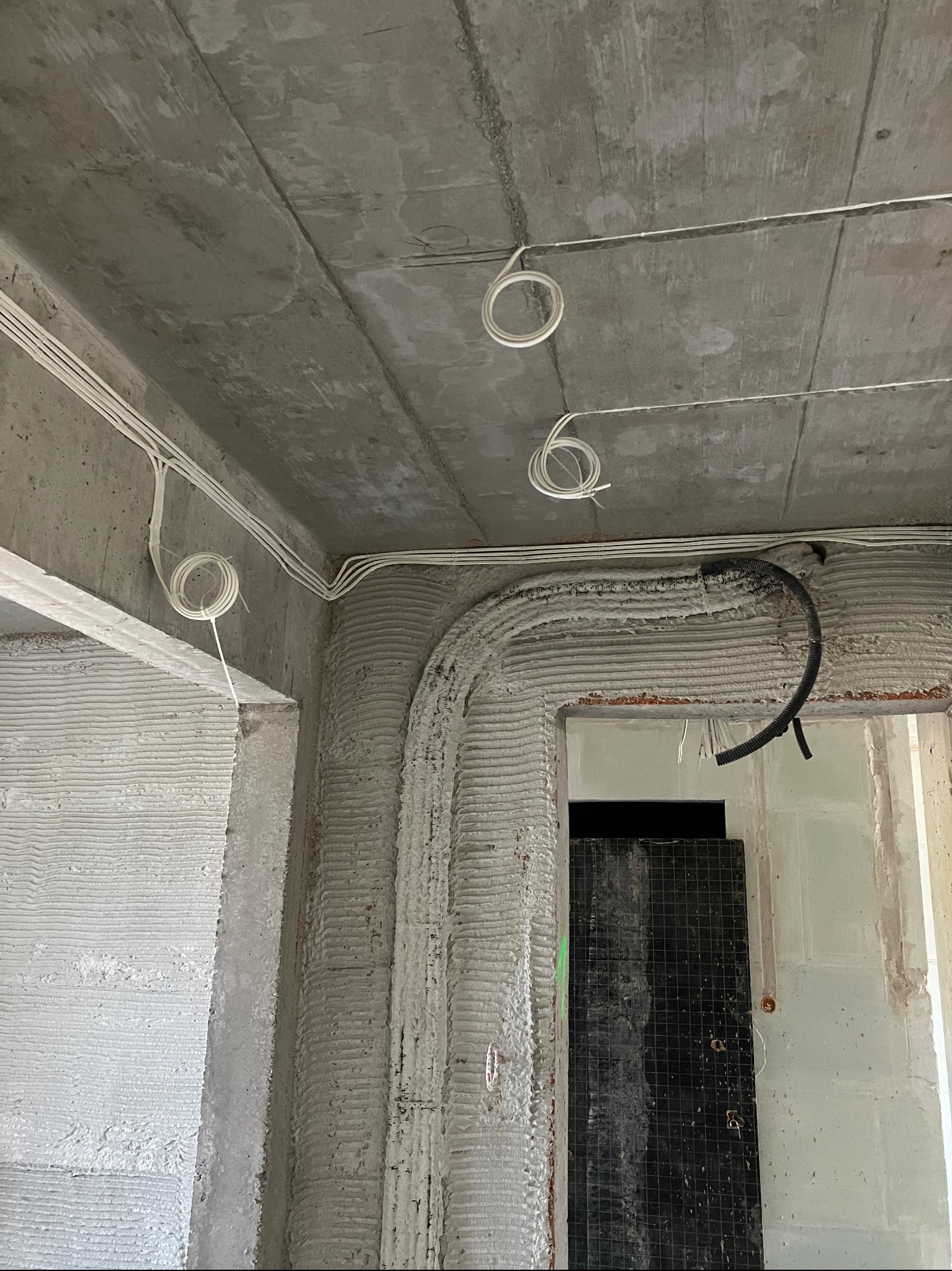 Surowy betonowy sufit i ściany w trakcie instalacji elektrycznej: widoczne białe kable elektryczne prowadzone po suficie i ścianach, częściowo przymocowane, z pętlami zwisającymi z sufitu,...