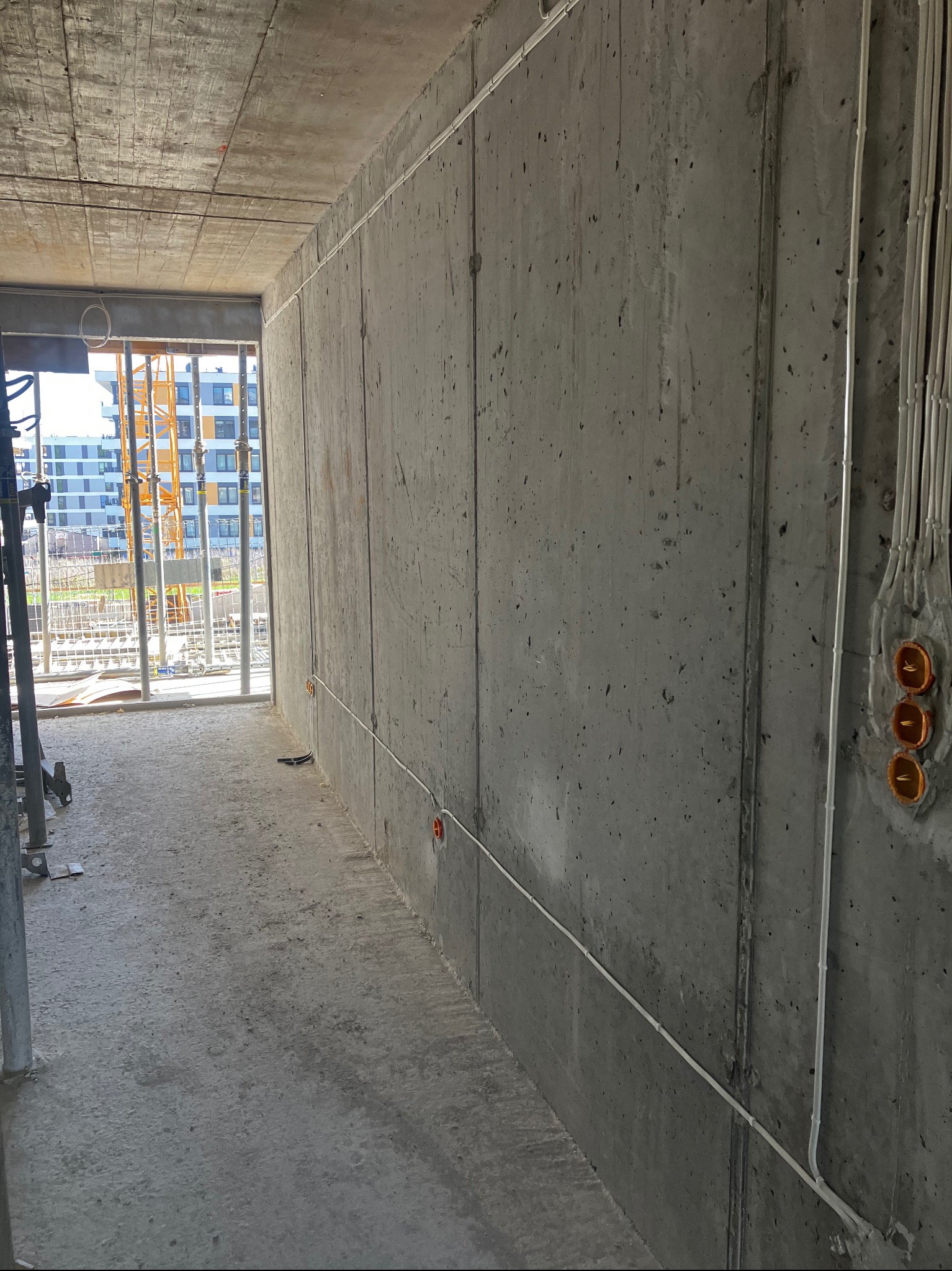 Surowy betonowy korytarz w nowym budynku z widocznymi przewodami elektrycznymi w białych rurkach, pomarańczowe puszki podtynkowe i widok na budowę za oknem.
