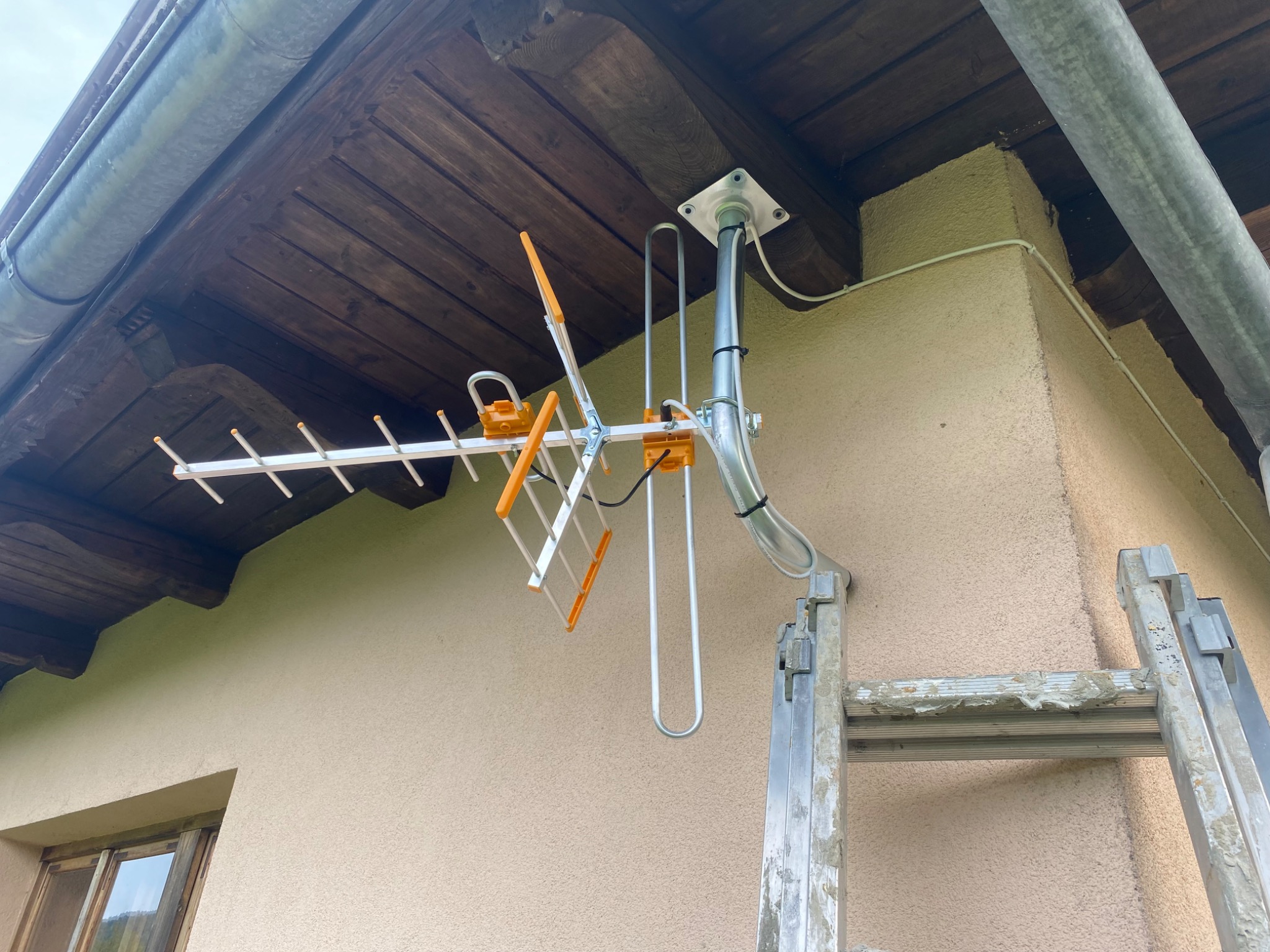 Zamontowana antena telewizyjna z pomarańczowymi elementami, przymocowana do ściany domu, widoczna drabina aluminiowa.