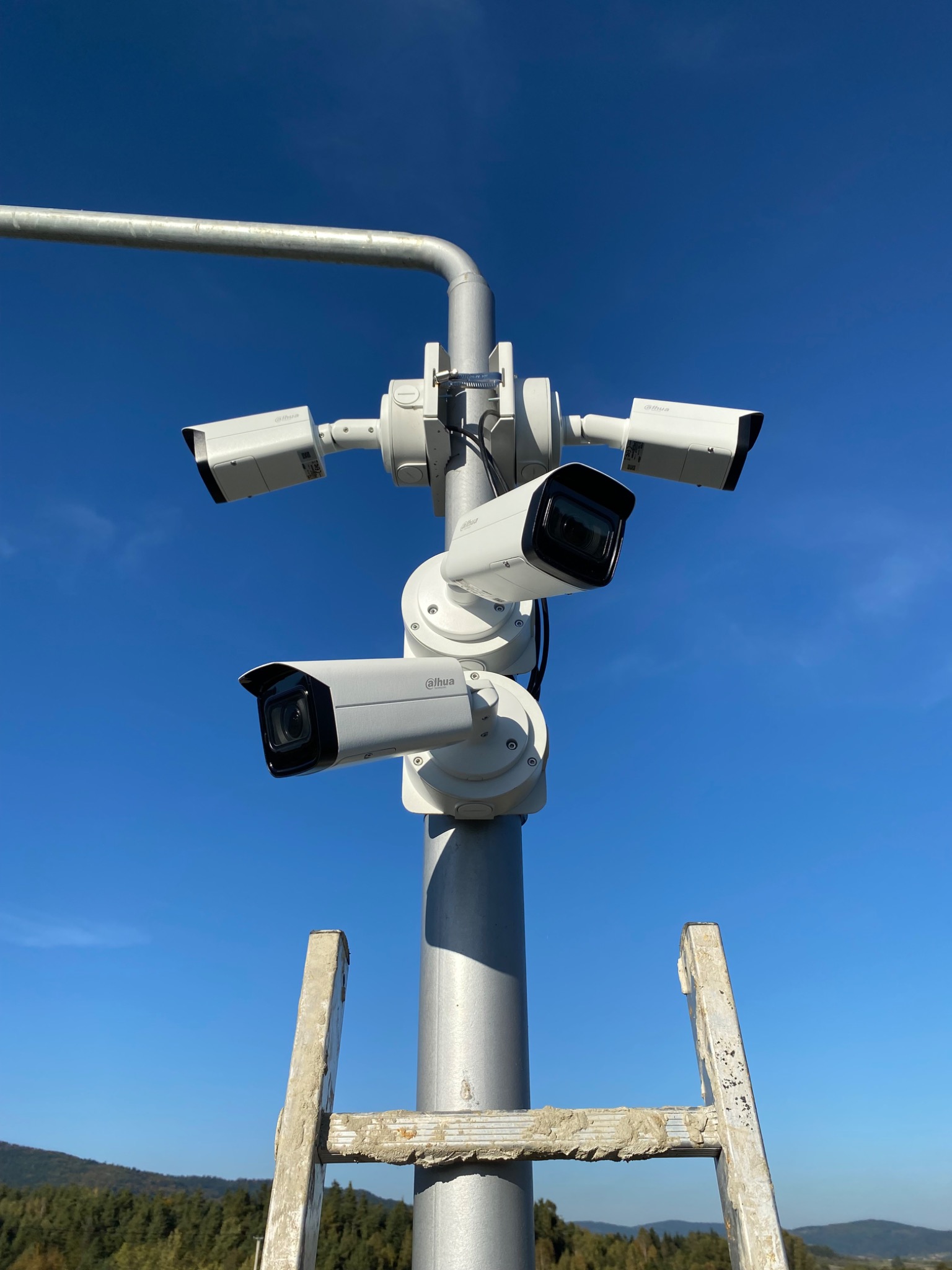 Zestaw czterech kamer monitoringu marki Dahua zamontowanych na metalowym słupie z drabinką w tle, widok z dołu na błękitne niebo i las.
