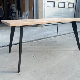 LoftStory Karol Miluski - Stół z jasnym blatem i czarnymi nogami w kształcie trapezu, dodatkowo wzmocniony cienkim metalowym prętem, ustawiony w warsztacie na betonowej posadzce.