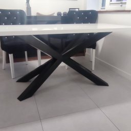 LoftStory Karol Miluski - Biały stół z czarnymi, skrzyżowanymi nogami w nowoczesnym wnętrzu, obok czarne krzesła z dekoracyjnymi guzikami, widoczne narzędzia na podłodze.