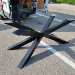 Czarna, metalowa podstawa stołu z ośmioma nogami, leżąca na parkingu, w tle biały samochód dostawczy z otwartymi drzwiami.