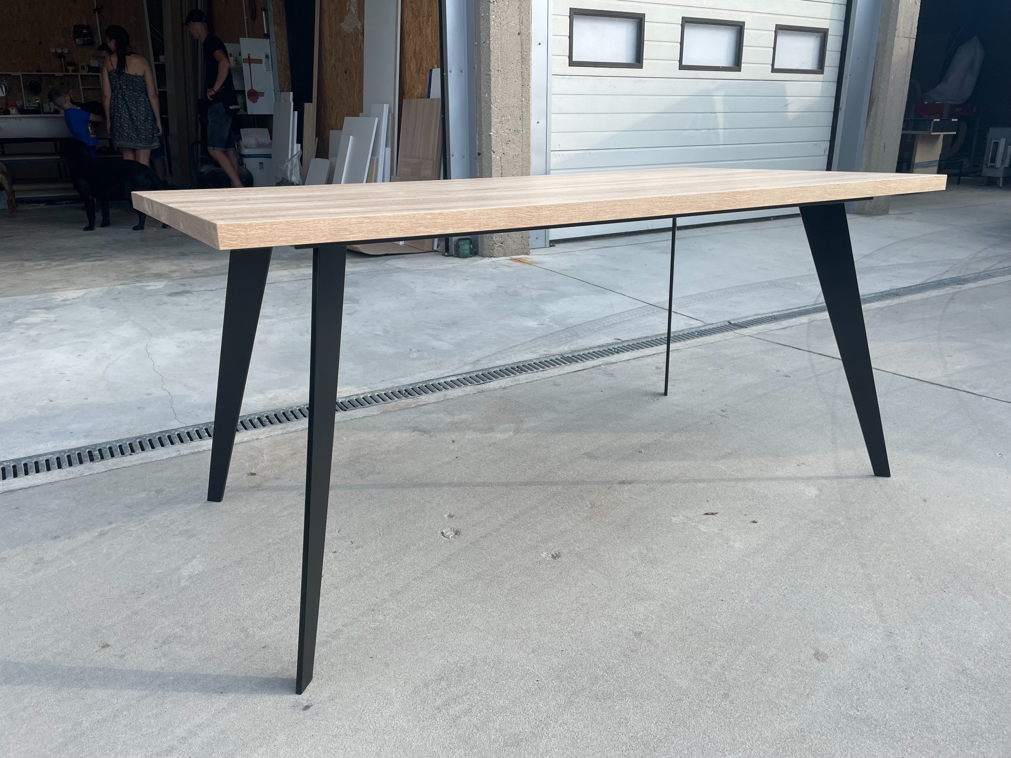 Stół z jasnym blatem i czarnymi nogami w kształcie trapezu, dodatkowo wzmocniony cienkim metalowym prętem, ustawiony w warsztacie na betonowej posadzce.