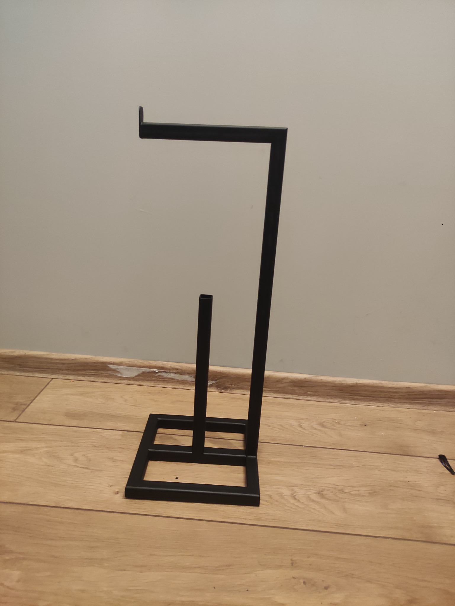 Czarny, metalowy stojak na papier toaletowy o minimalistycznym, geometrycznym designie, ustawiony na drewnianej podłodze przy szarej ścianie.