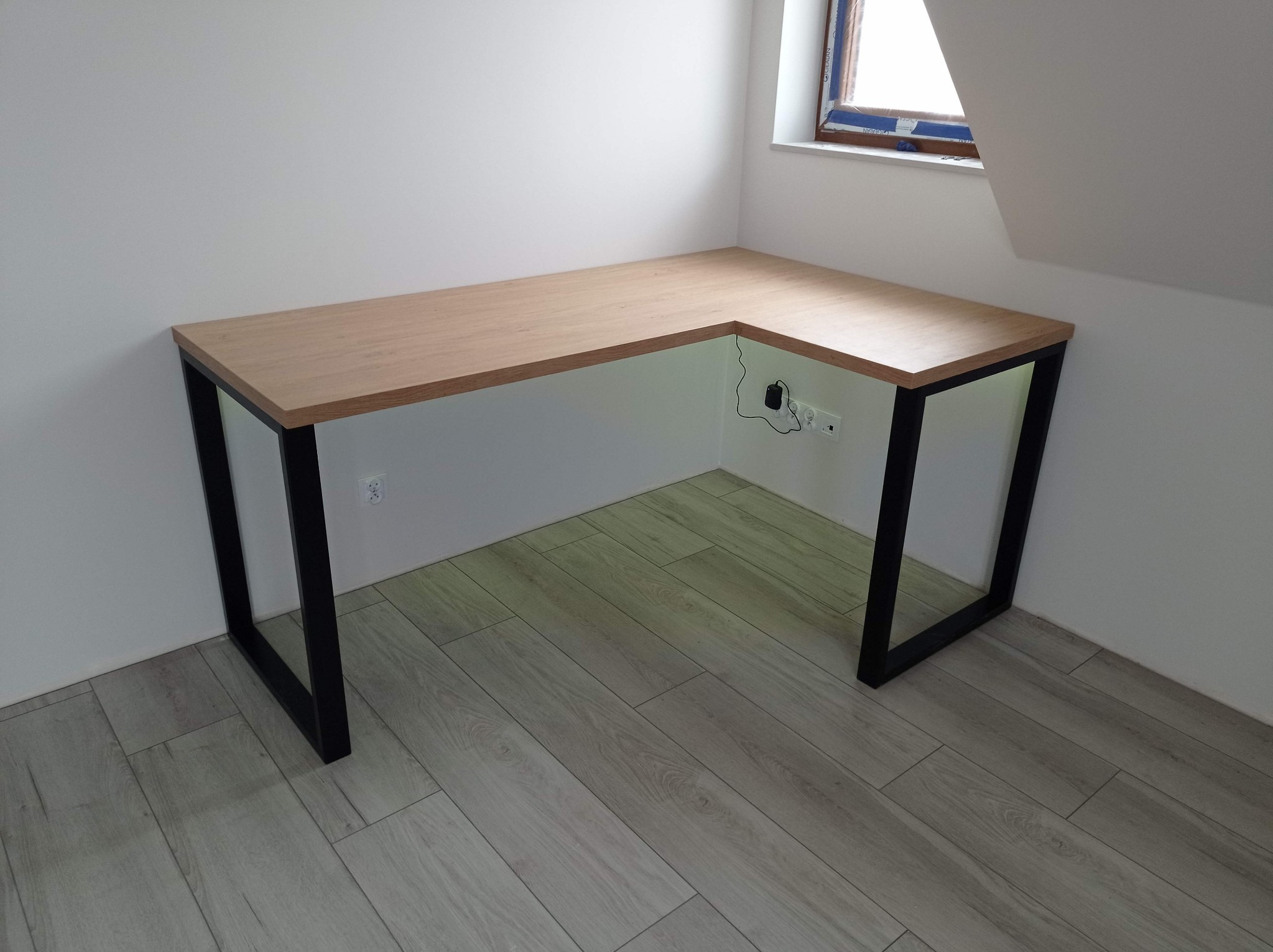 Narożne biurko z drewnianym blatem i czarnymi metalowymi nogami w minimalistycznym wnętrzu z drewnianą podłogą i oknem na poddaszu.