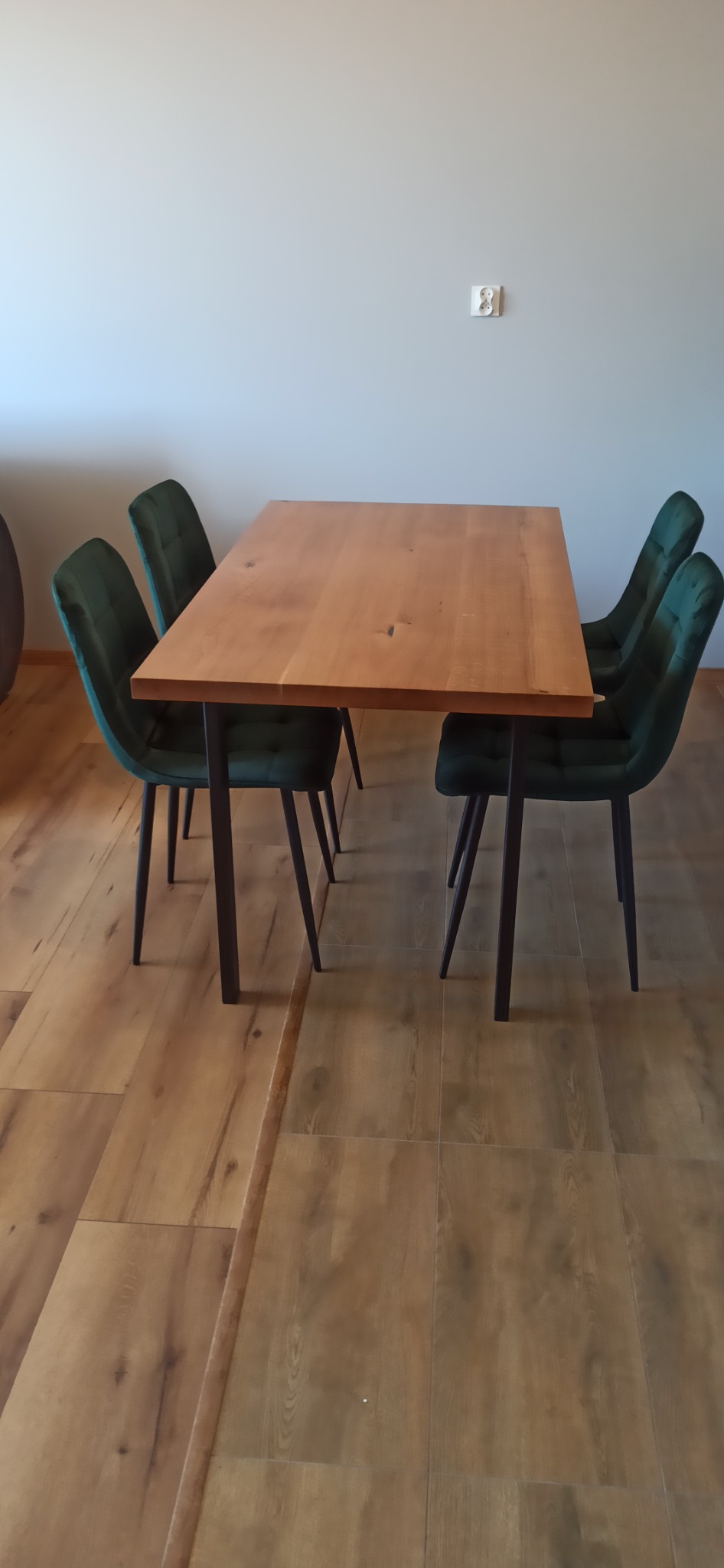 Prostokątny stół z blatem z jasnego drewna i ciemnymi, metalowymi nogami, otoczony czterema krzesłami z ciemnozieloną tapicerką welurową, ustawiony na podłodze imitującej drewno.