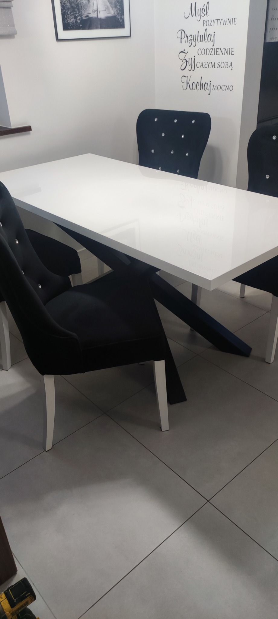 Nowoczesny stół z białym blatem i czarnymi nogami, otoczony czarnymi krzesłami z kryształkami, na tle ściany z napisem 'Myśl pozytywnie, Przytulaj codziennie, Żyj całym sobą, Kochaj mocno'.