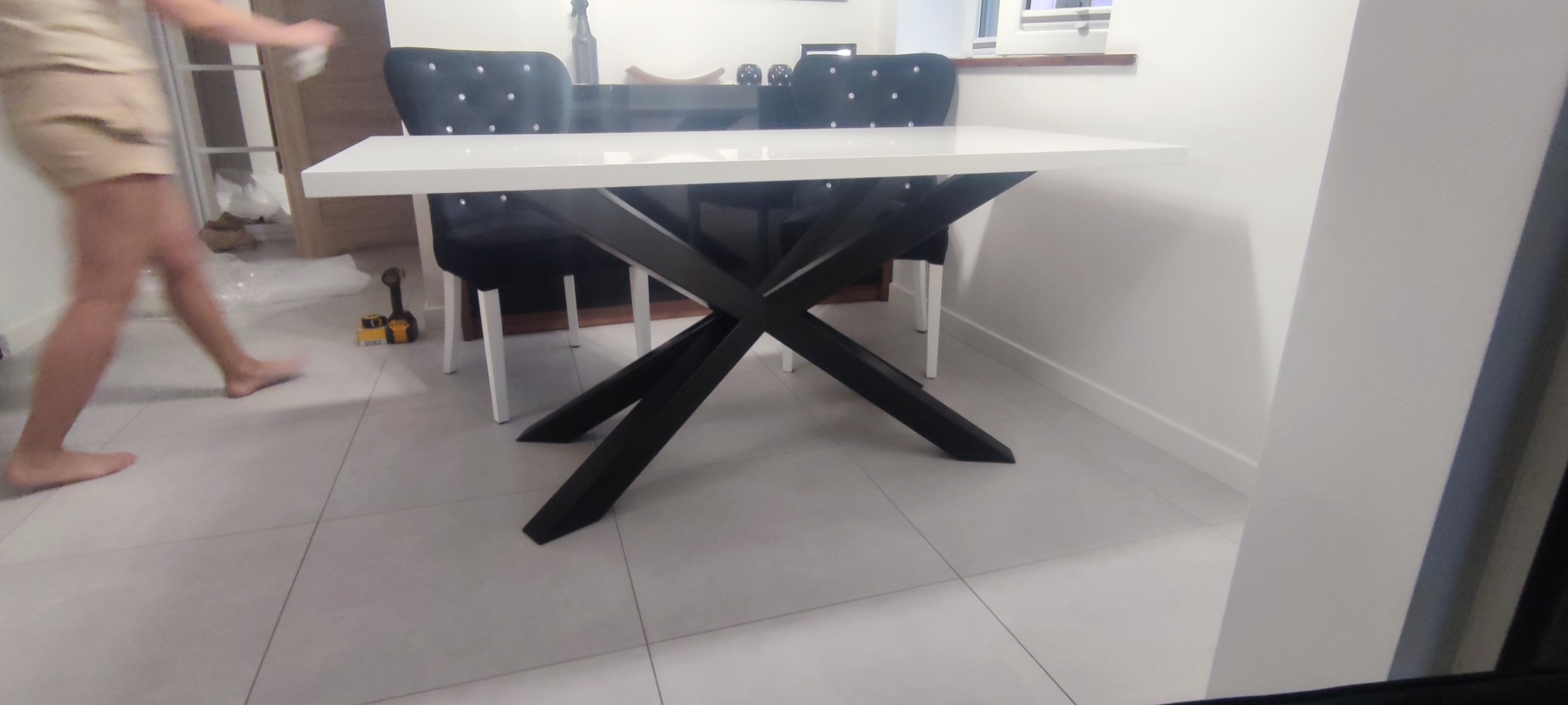 Biały stół z czarnymi, skrzyżowanymi nogami w nowoczesnym wnętrzu, obok czarne krzesła z dekoracyjnymi guzikami, widoczne narzędzia na podłodze.