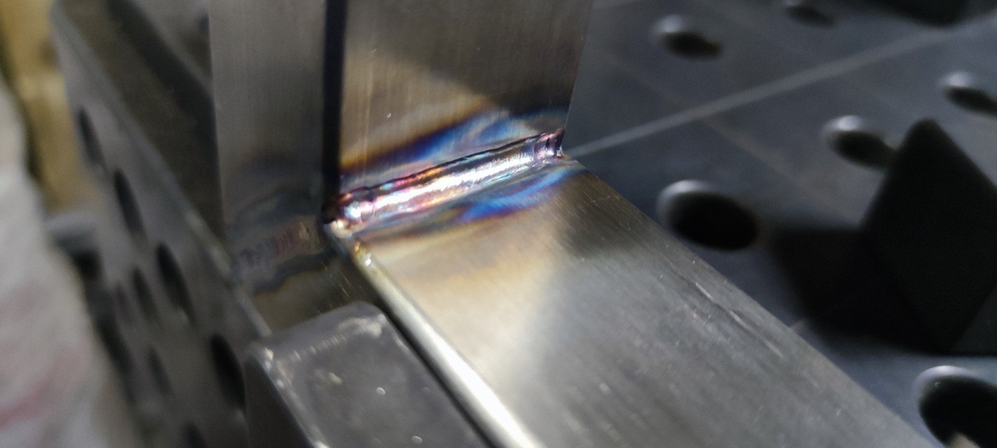 Szlifowany profil stalowy po spawaniu, widoczne tęczowe przebarwienia termiczne w okolicy spoiny, ułożony na perforowanym stole spawalniczym.