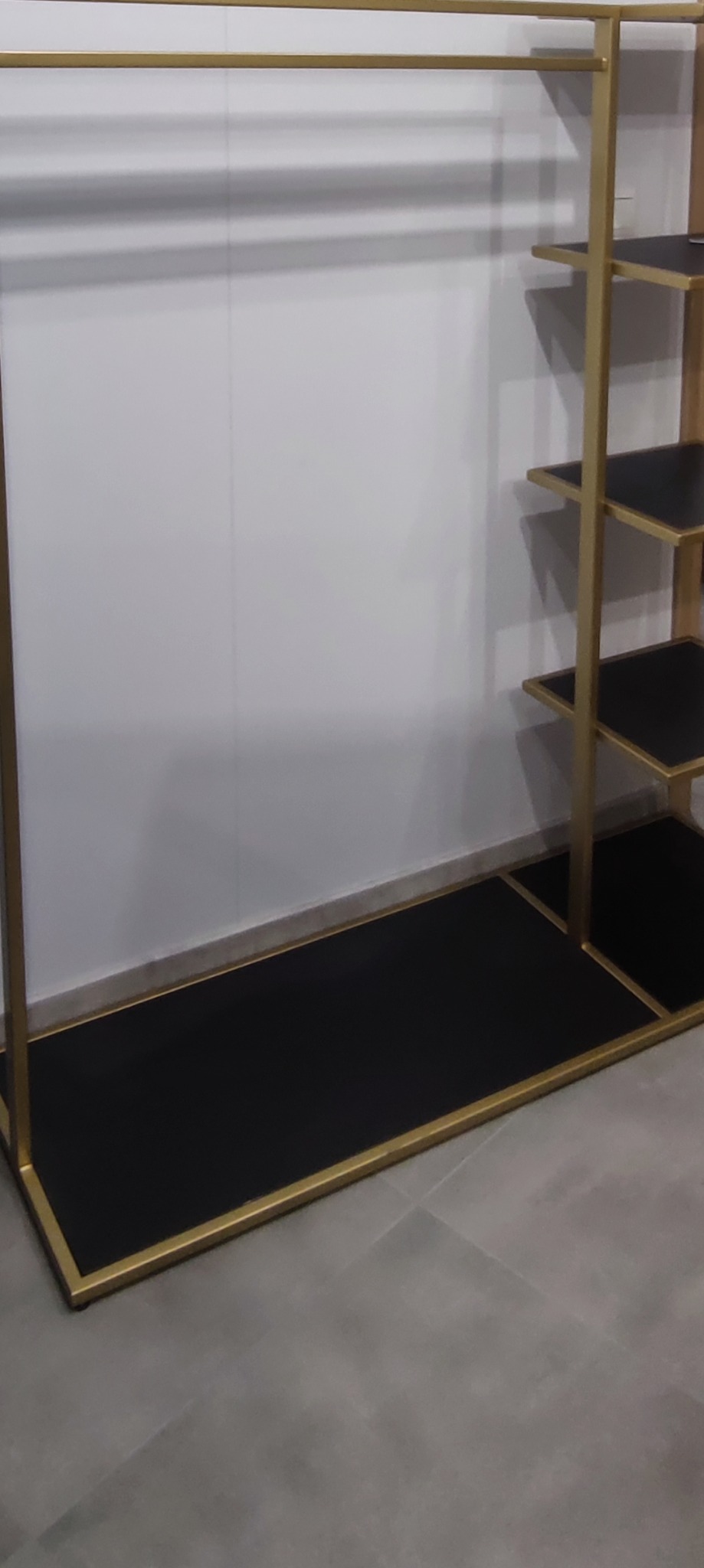 Metalowy regał z półkami i drążkiem na ubrania, pomalowany na złoto i czarno, stojący przy białej ścianie.