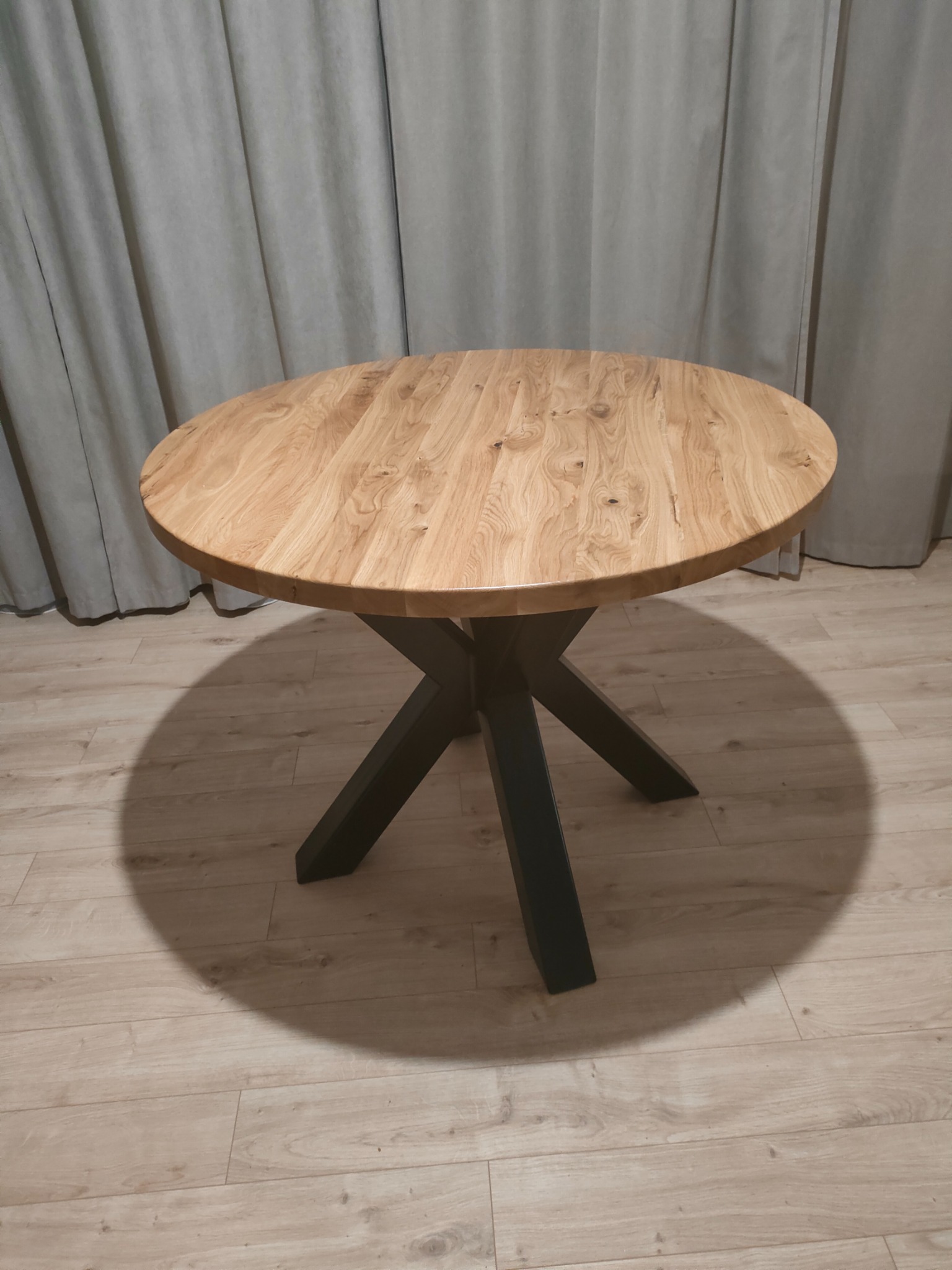 Okrągły stół z blatem z jasnego drewna i czarną, metalową podstawą w kształcie gwiazdy, wykonany w Łapach, na tle szarych zasłon i drewnianej podłogi.
