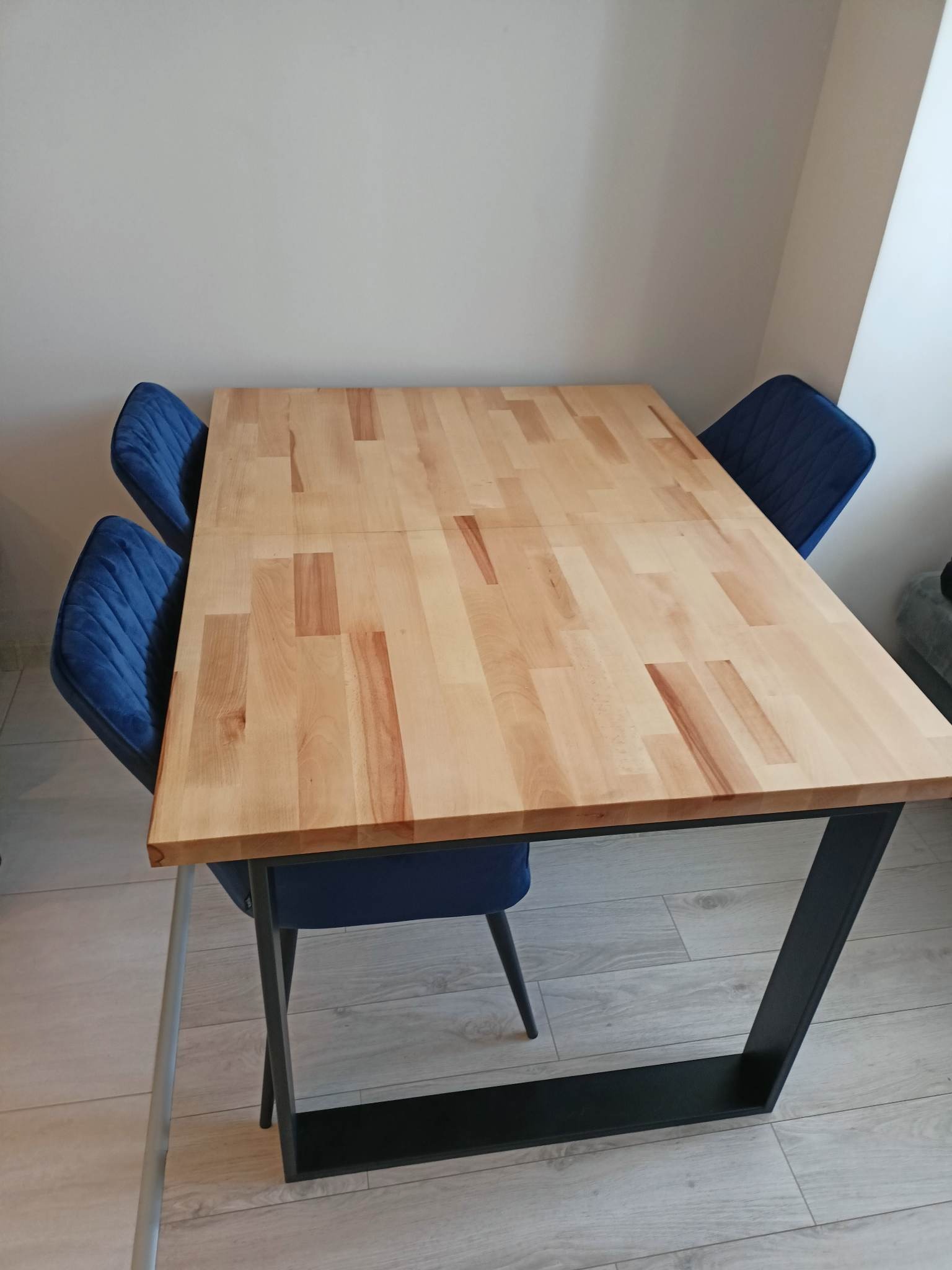 Rozkładany stół z blatem z jasnego drewna i czarną, metalową podstawą, otoczony trzema krzesłami z niebieską tapicerką.