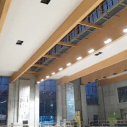 Wnętrze hali w trakcie wykończenia, widoczne drewniane belki stropowe z oświetleniem i częściowo zamontowane białe panele sufitowe, duże okna i betonowe ściany.