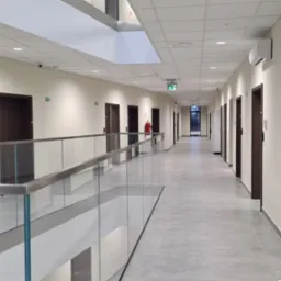 Długi korytarz biurowy z szarymi płytkami podłogowymi, białymi ścianami, ciemnobrązowymi drzwiami i szklaną balustradą, oświetlony lampami sufitowymi i z widocznymi elementami wentylacji...