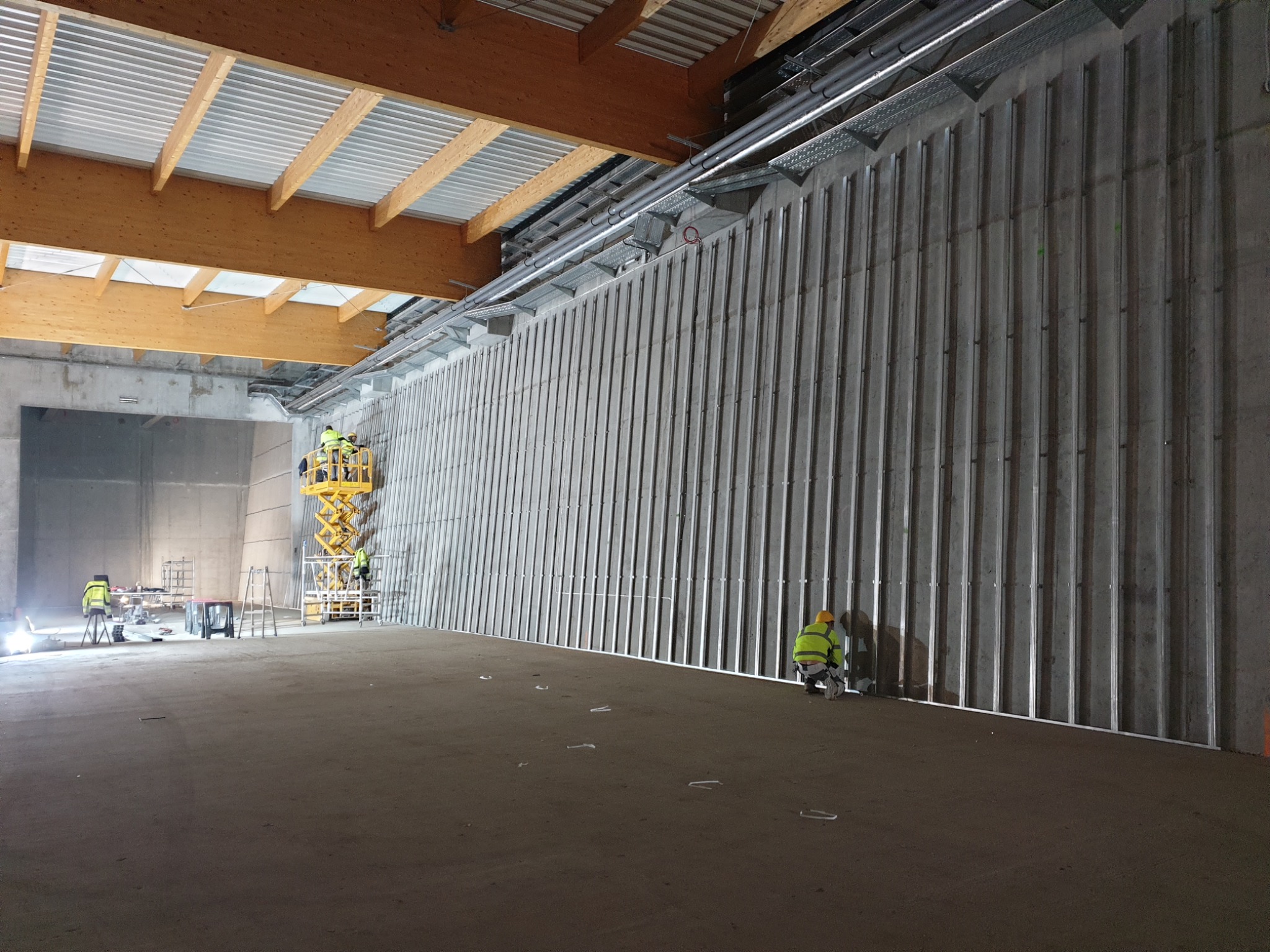 Montaż metalowej konstrukcji na betonowej ścianie w hali. Pracownicy na podnośniku i przy ścianie. Widoczne elementy instalacji pod sufitem. Surowy stan wykończenia.