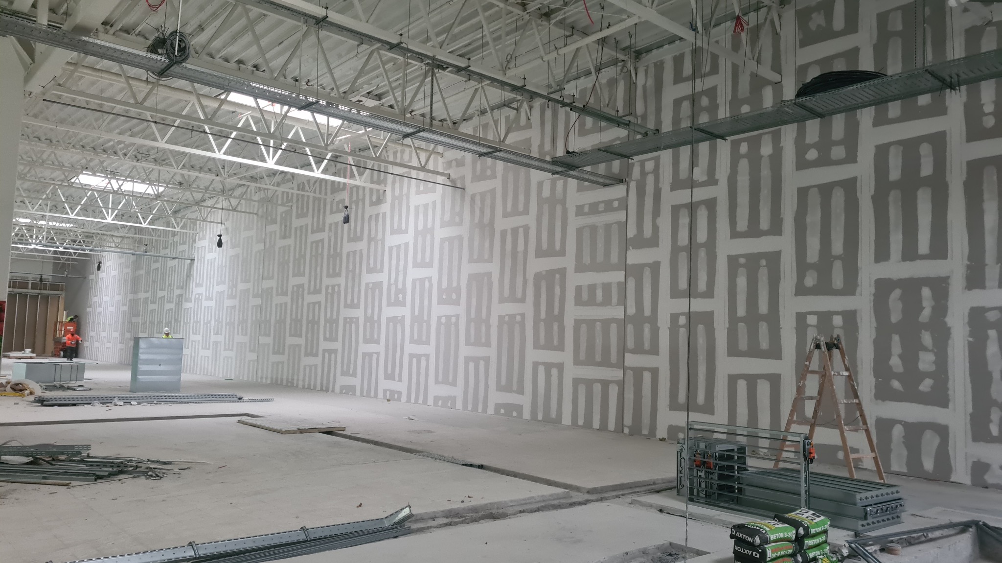 Szeroki kadr na wnętrze w trakcie prac wykończeniowych: ściany z płyt gipsowo-kartonowych z widocznymi łączeniami, metalowa konstrukcja sufitu, drabina i materiały budowlane.