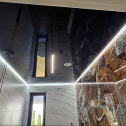 Wnętrze łazienki z czarnym, lustrzanym sufitem, oświetleniem LED wzdłuż krawędzi sufitu i ścian, oraz marmurowymi płytkami w odcieniach bieli i brązu na ścianach; widoczne okno.