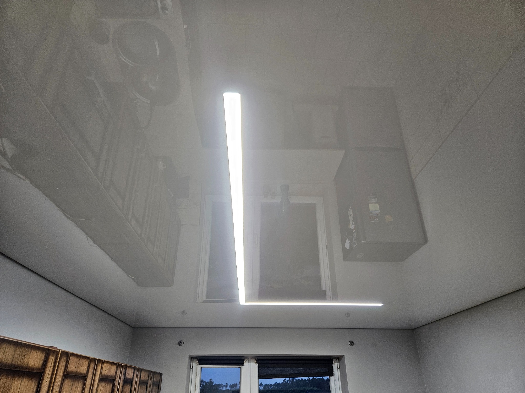 Biały, błyszczący sufit z odbiciem wyposażenia kuchni i oświetleniem LED w kształcie litery L. Widok na okno z roletami i fragment szafek kuchennych.