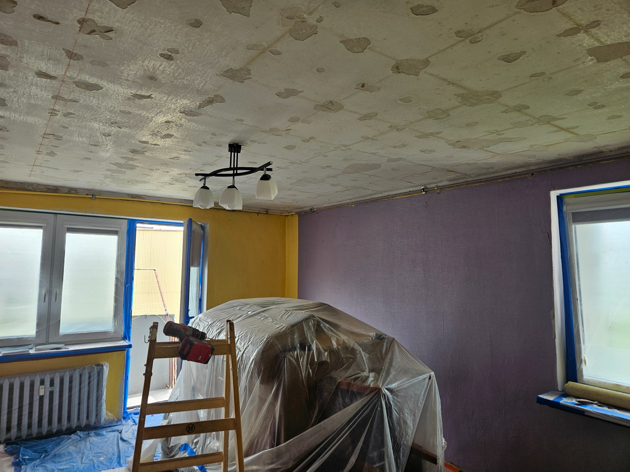 Przygotowanie do remontu: Pokój z uszkodzonym sufitem, meble zabezpieczone folią, drabina z wiertarką, okna i grzejnik oklejone taśmą malarską, ściany w kolorze żółtym i fioletowym.