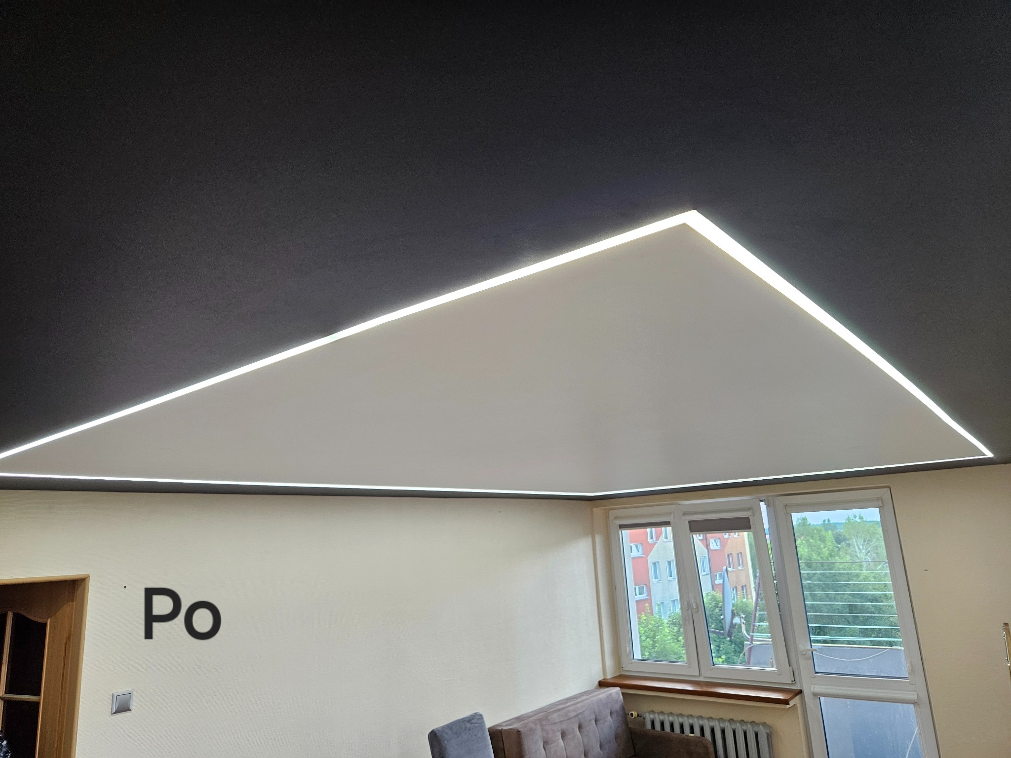 Nowoczesny salon z czarnym sufitem i trójkątnym białym obniżeniem z oświetleniem LED, widok na okno i napis 'Po' na ścianie.