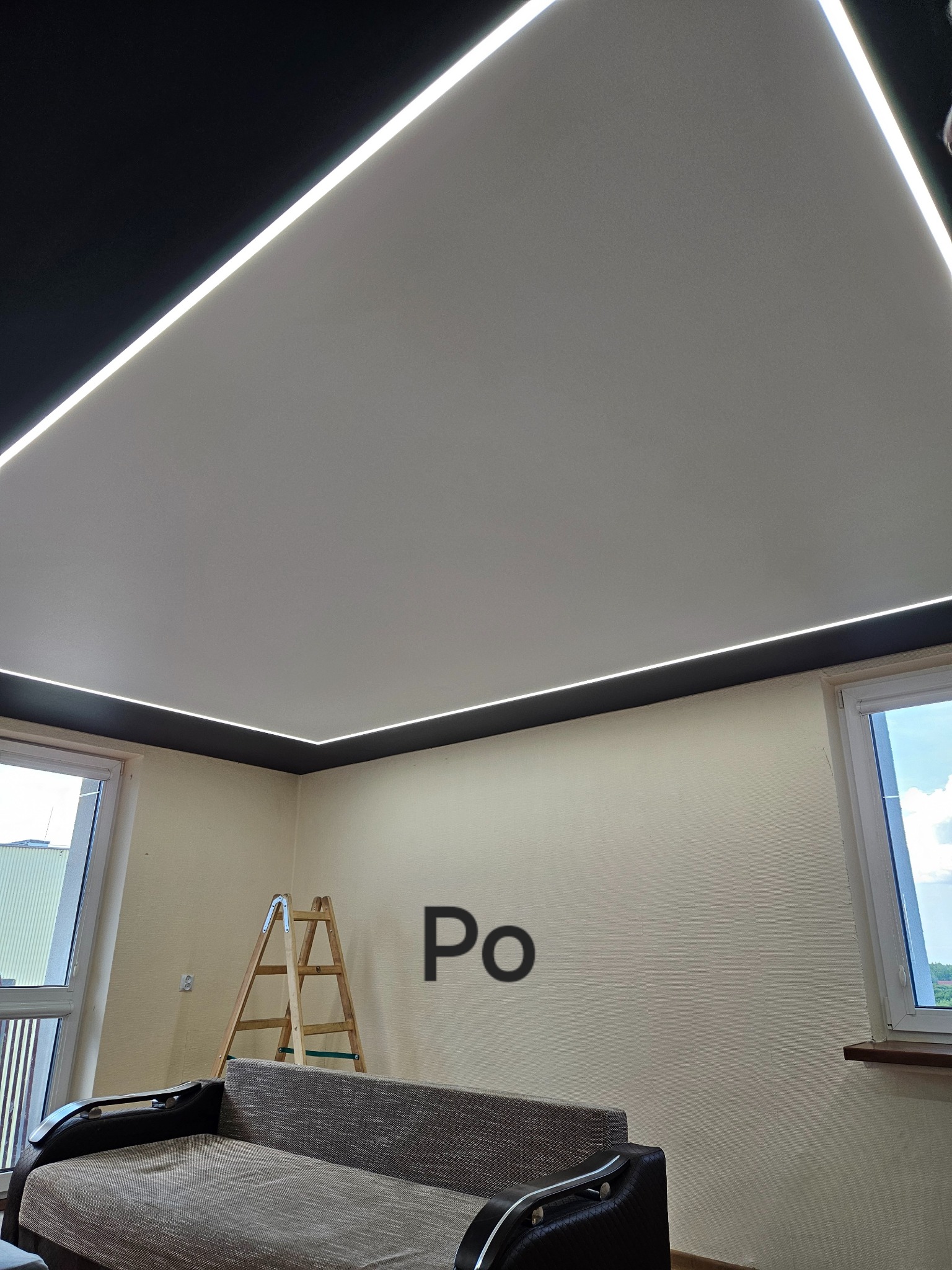 Jasny, gładki sufit z wbudowanym oświetleniem LED w kształcie prostokąta, czarna listwa sufitowa, drabina oparta o ścianę, widok z dołu, sofa w pomieszczeniu.