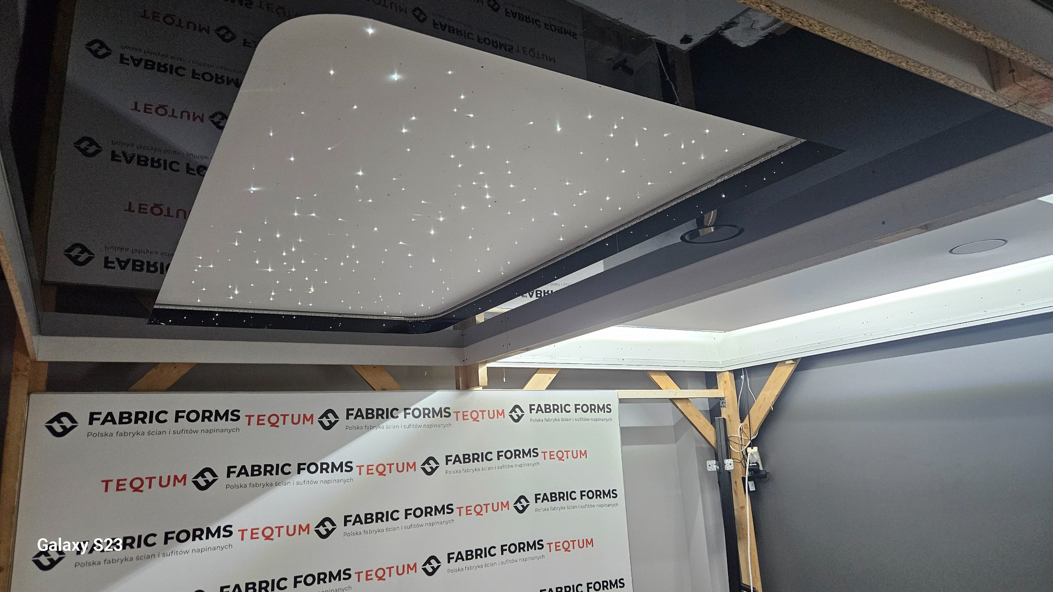 Montaż sufitu napinanego z efektem rozgwieżdżonego nieba, widoczne elementy konstrukcyjne i baner reklamowy firmy.