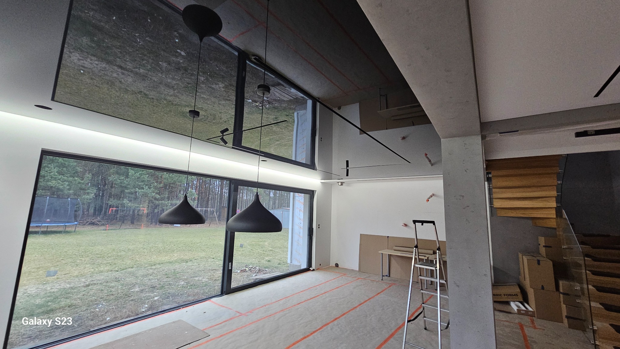 Wnętrze w trakcie prac wykończeniowych: widoczne duże okna z widokiem na zewnątrz, sufit z elementami czarnego szkła, minimalistyczne lampy wiszące, ślady po taśmie malarskiej na podłodze, drabina...