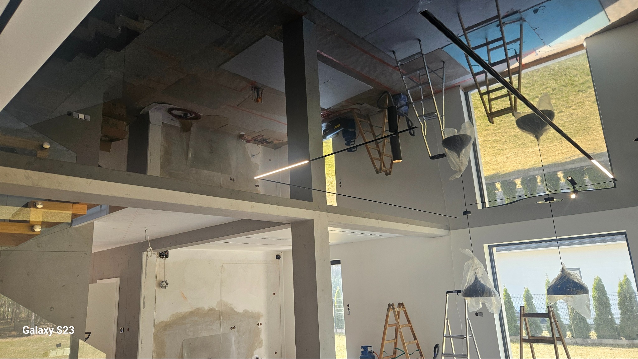 Wnętrze w trakcie wykańczania: widoczne schody z drewnianymi stopniami, surowy beton, ściany przygotowywane do malowania, drabiny, lampy wiszące zabezpieczone folią, odblaskowe sufity.