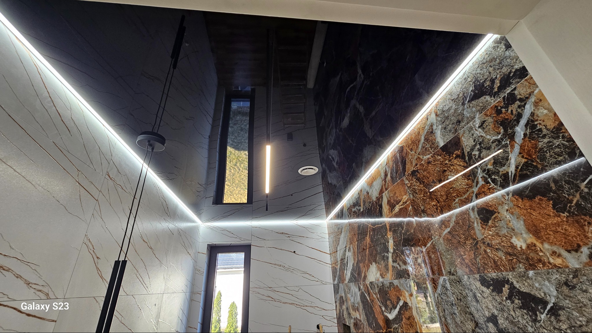 Wnętrze łazienki z czarnym, lustrzanym sufitem, oświetleniem LED wzdłuż krawędzi sufitu i ścian, oraz marmurowymi płytkami w odcieniach bieli i brązu na ścianach; widoczne okno.